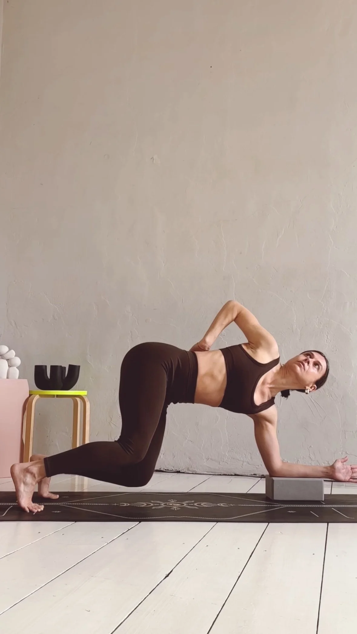 A woman performing a yoga pose on a black mat in a room with a beige wall. She is in a modified side plank position, supporting herself with one forearm on a yoga block, one knee on the mat, and the other leg extended back. She is wearing a black sports bra and black leggings.