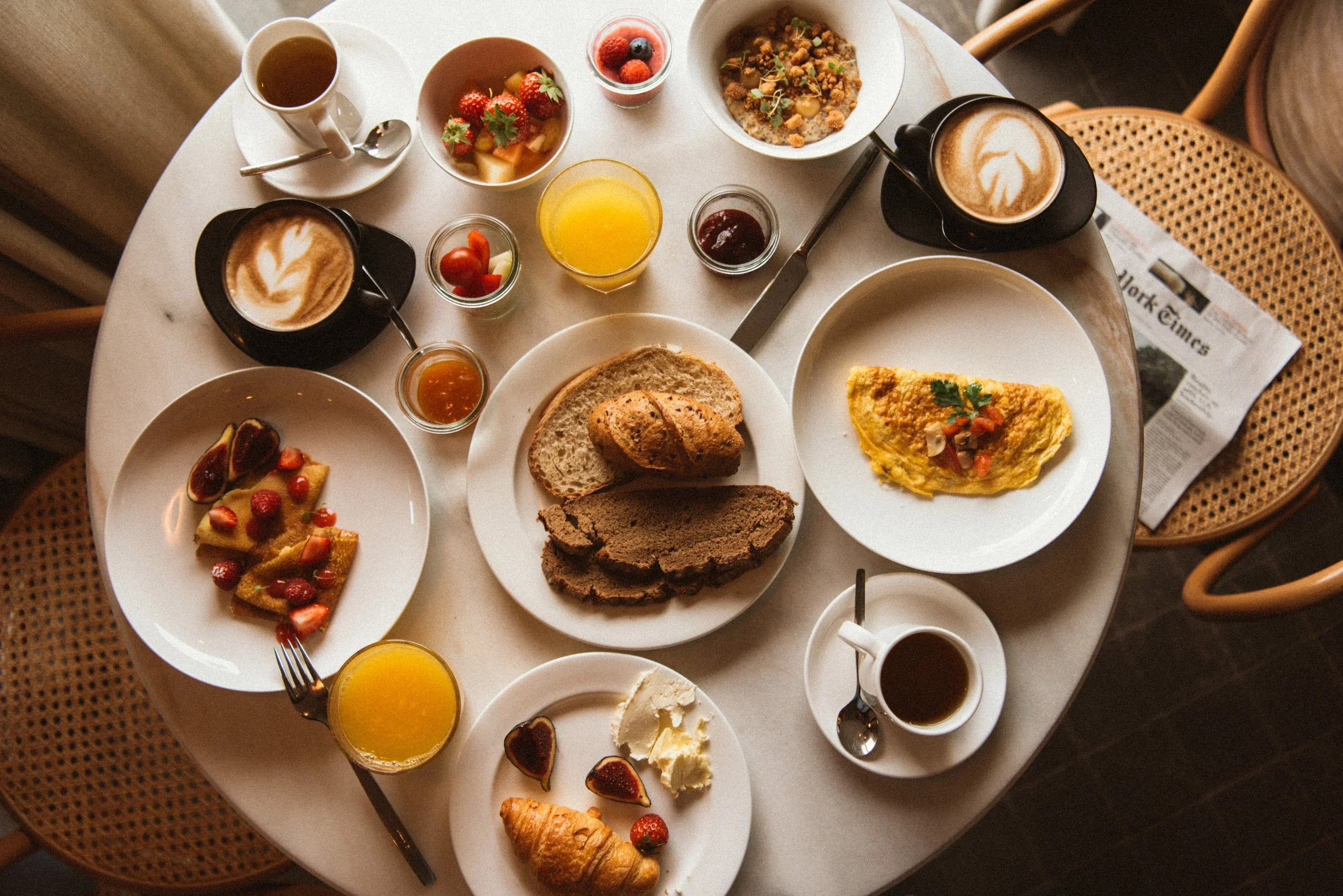 A breakfast spread on a white marble table includes croissants, bread, a variety of fruits like strawberries, figs, and raspberries, bowls of yogurt with strawberries, a small glass of orange juice, cups of coffee with latte art, a plate with an omelet, and slices of cake or bread, alongside a newspaper on a woven chair.