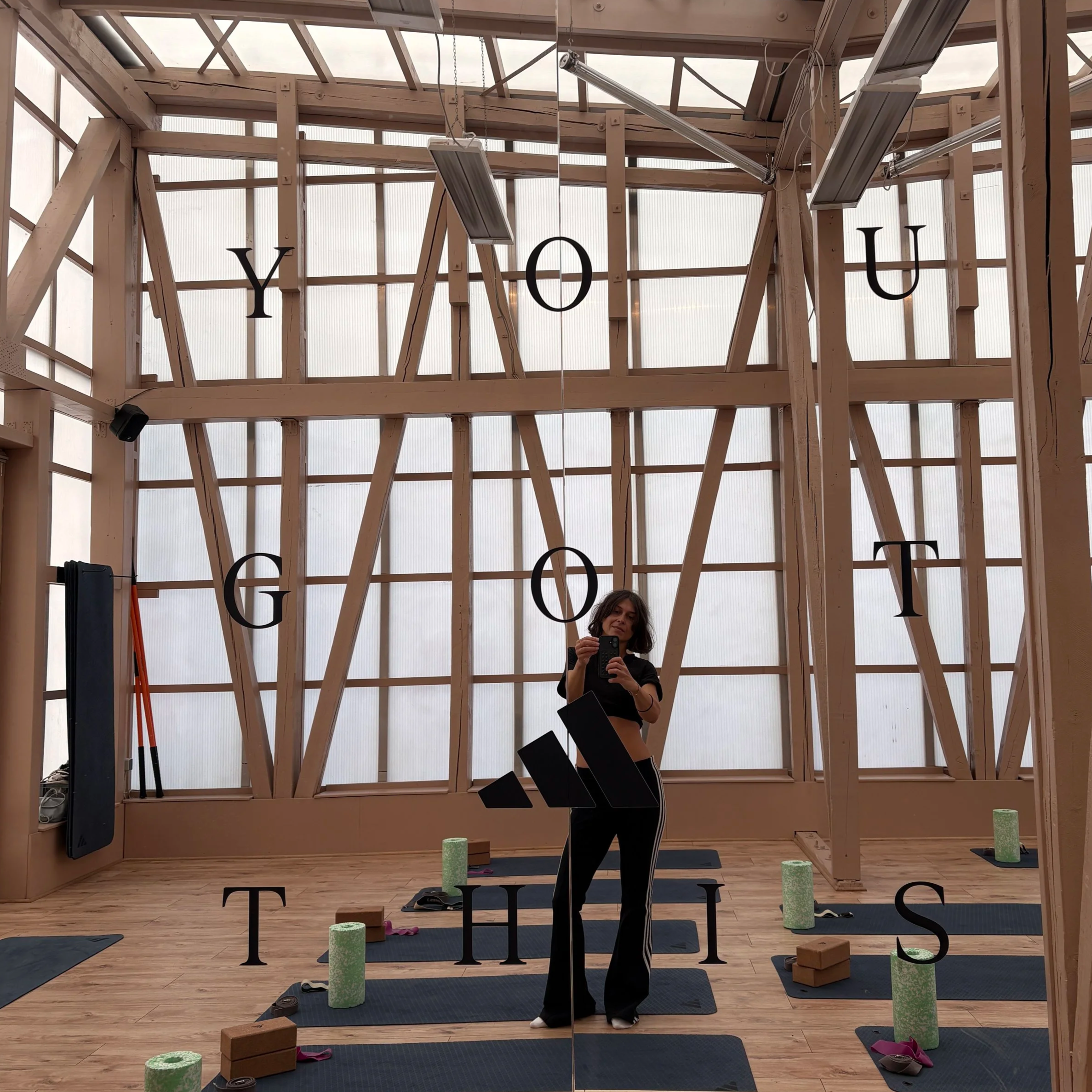 Woman in workout attire taking a selfie in a yoga studio with an unfinished wooden wall and yoga mats on the floor. The mirrored wall has the words 'YOGA' and 'THIS' arranged around her.