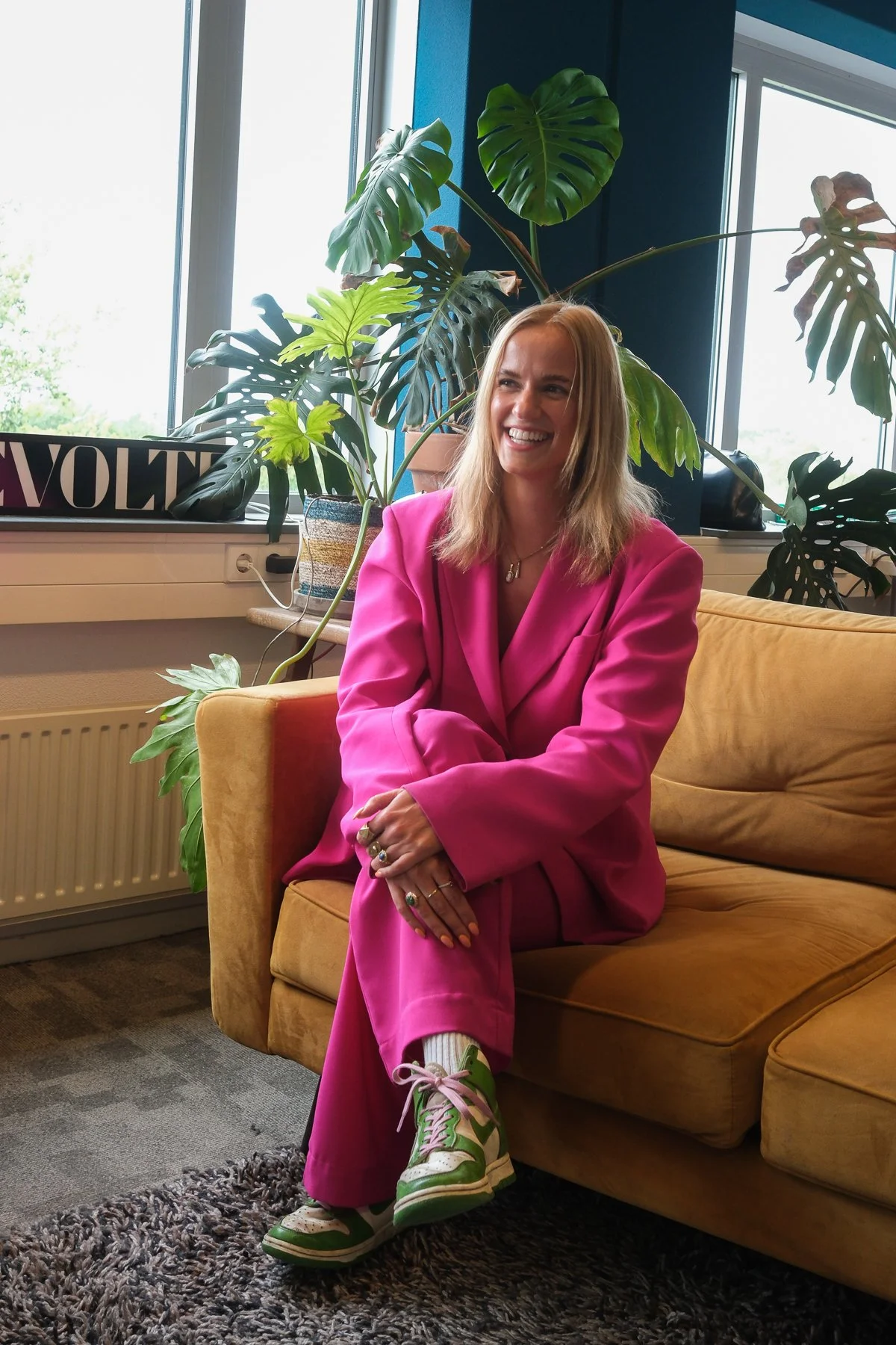 A woman with blonde hair smiling, wearing a bright pink suit, sitting on a yellow armchair in a room with large window and green plants, including a monstera plant behind her, with sunlight illuminating the space.