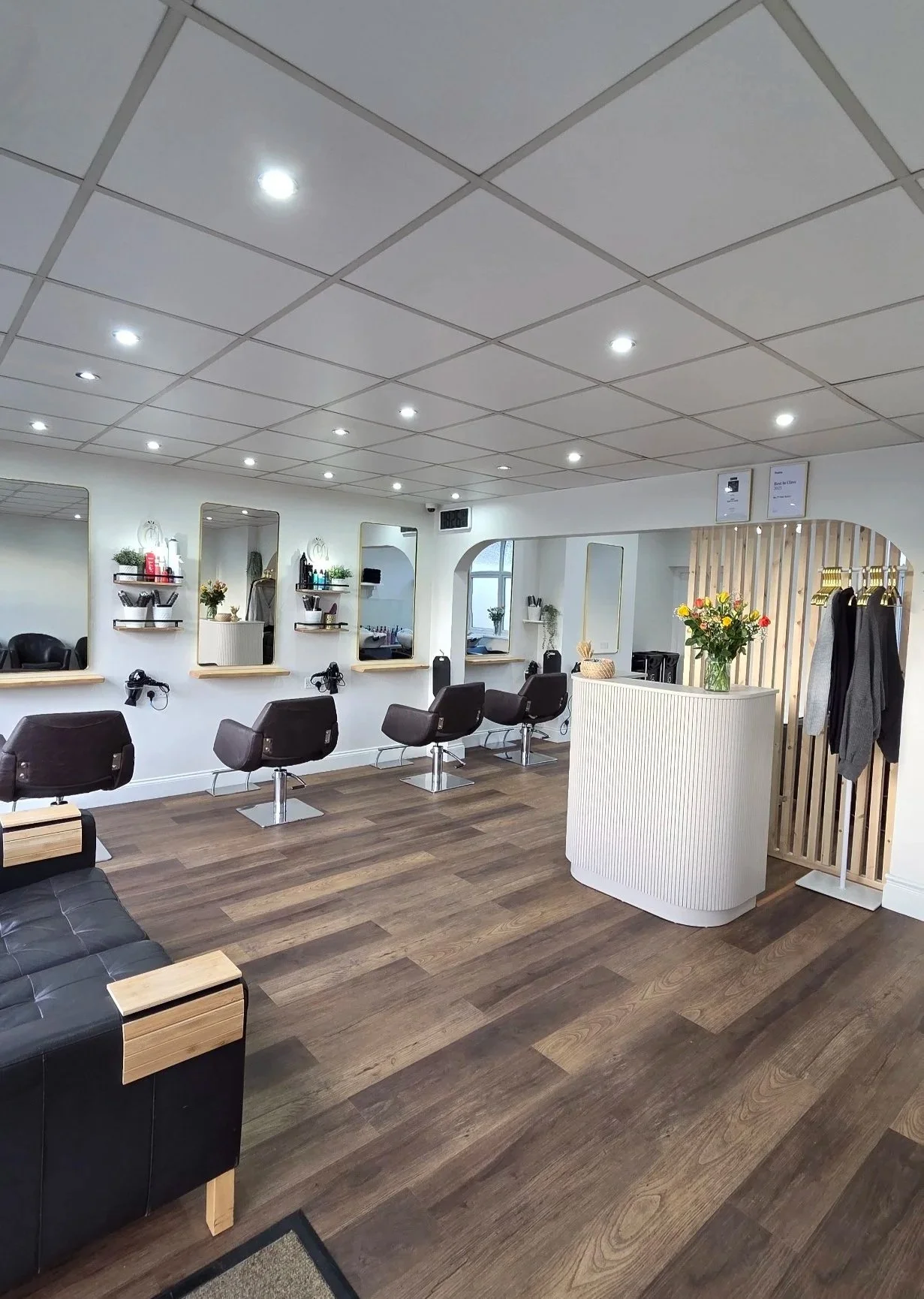 Interior of a modern hair salon with four styling chairs in front of mirrors, a reception desk with a vase of flowers, and wooden floors.