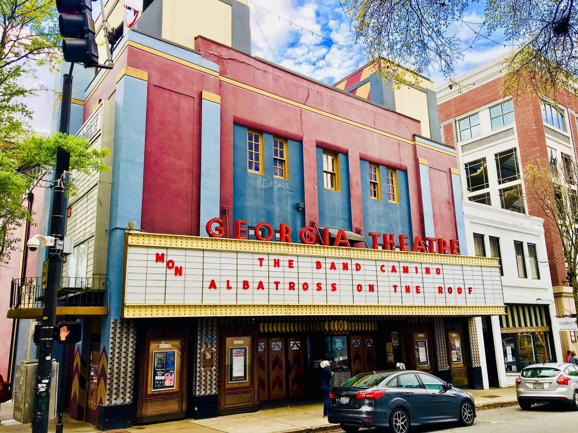 A colorful, vintage-style theater with a large marquee displaying "GEORGIA THEATRE" in red letters. The marquee lists upcoming performances: "THE BAND CAMINO" and "ALBATROSS ON THE ROOF."