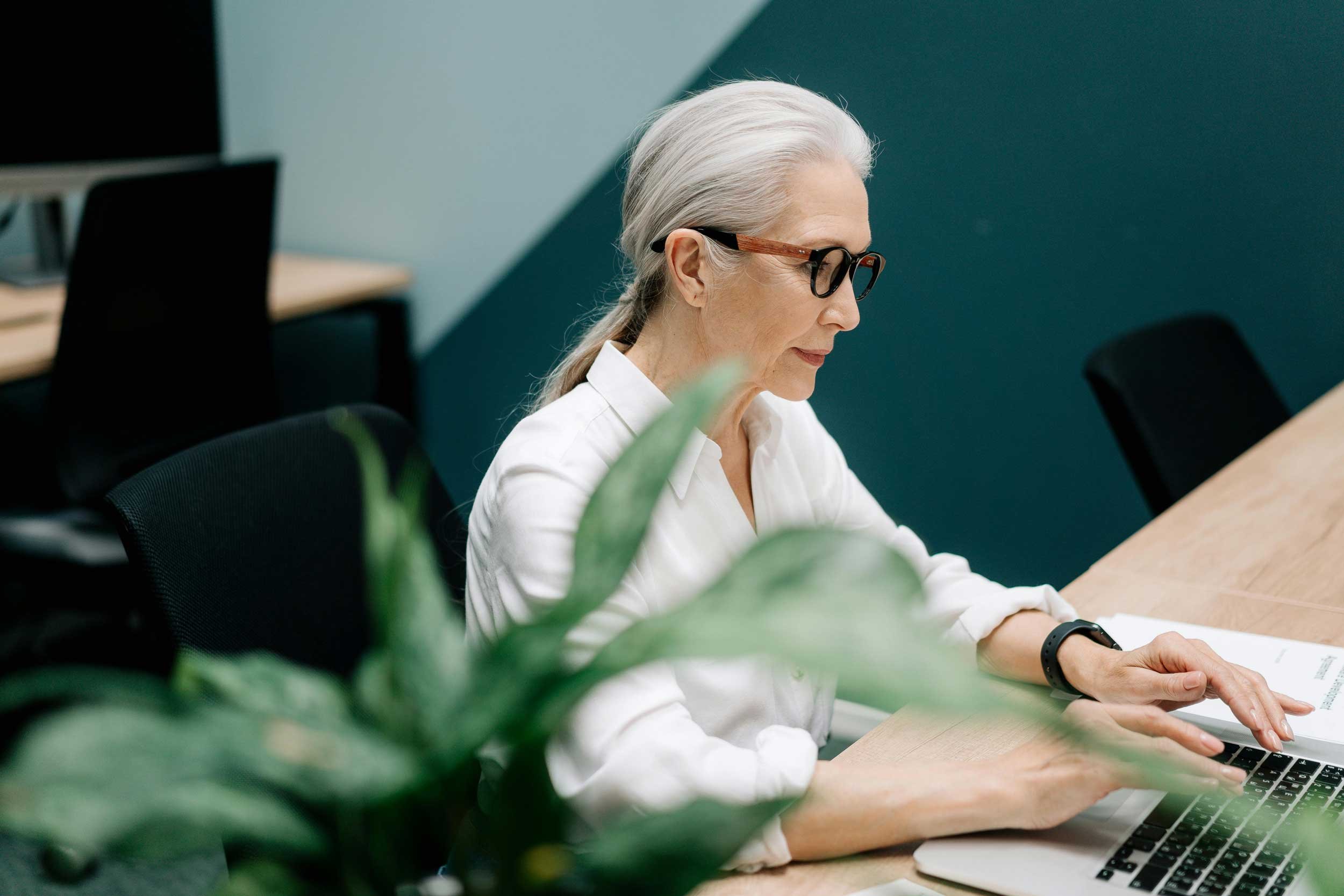 An older woman with gray hair in a ponytail sits at a desk in a modern office, typing on a laptop.