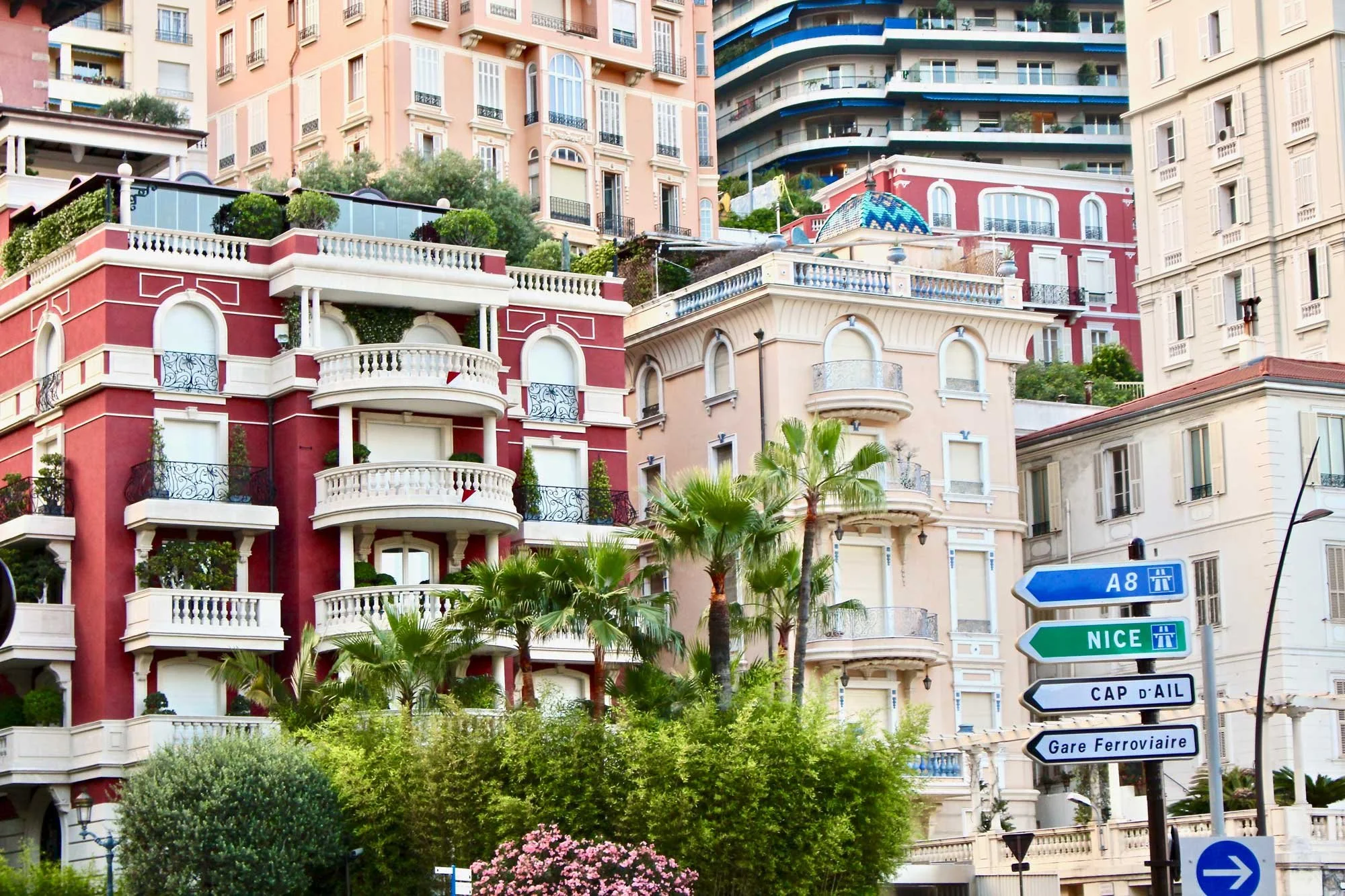 Colorful, multi-story buildings with balconies line a hillside. A road sign indicates directions to Nice, the A8 highway, and the railway station.