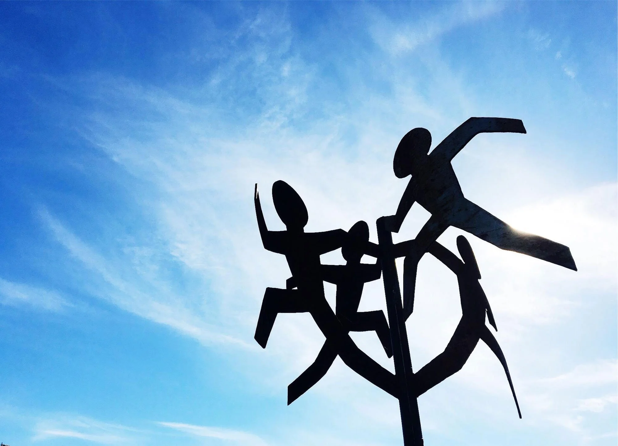 A silhouette of a metal sculpture features multiple human figures in dynamic, running poses, set against a bright blue sky with wispy clouds