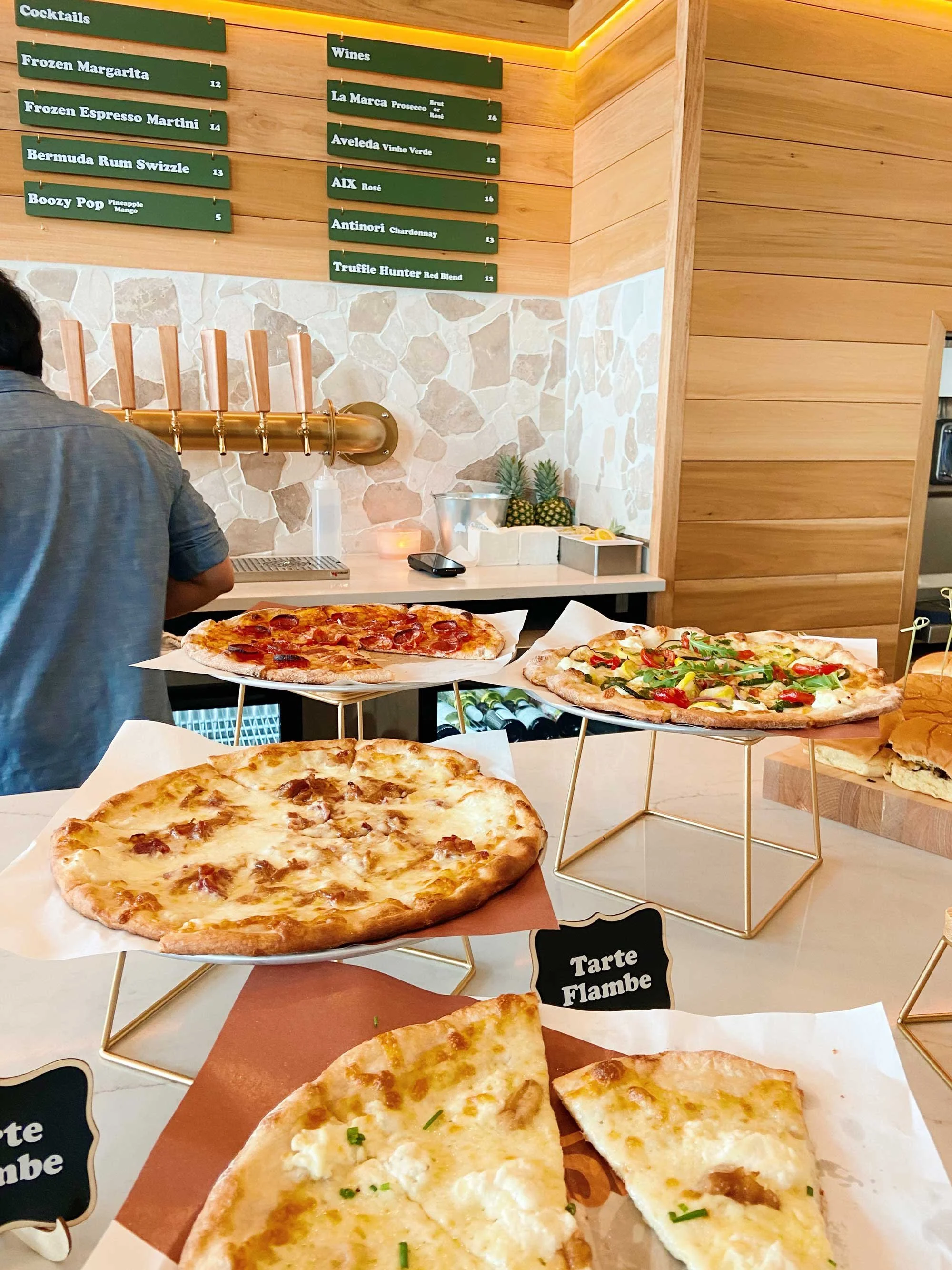 Display of various pizzas on stands. A person stands at a counter in the background, with menu boards listing cocktails and wines above.