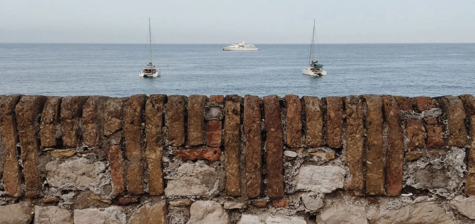 A stone wall partially obscures a view of a calm ocean. Two smaller sailboats are closer to the wall, while a large yacht is further out