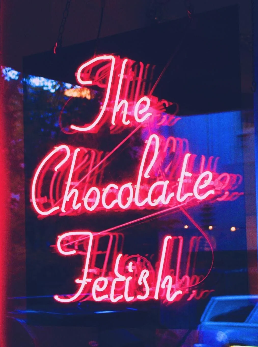 Glowing pink neon sign reading "The Chocolate Fetish" mounted against a dark, reflective surface