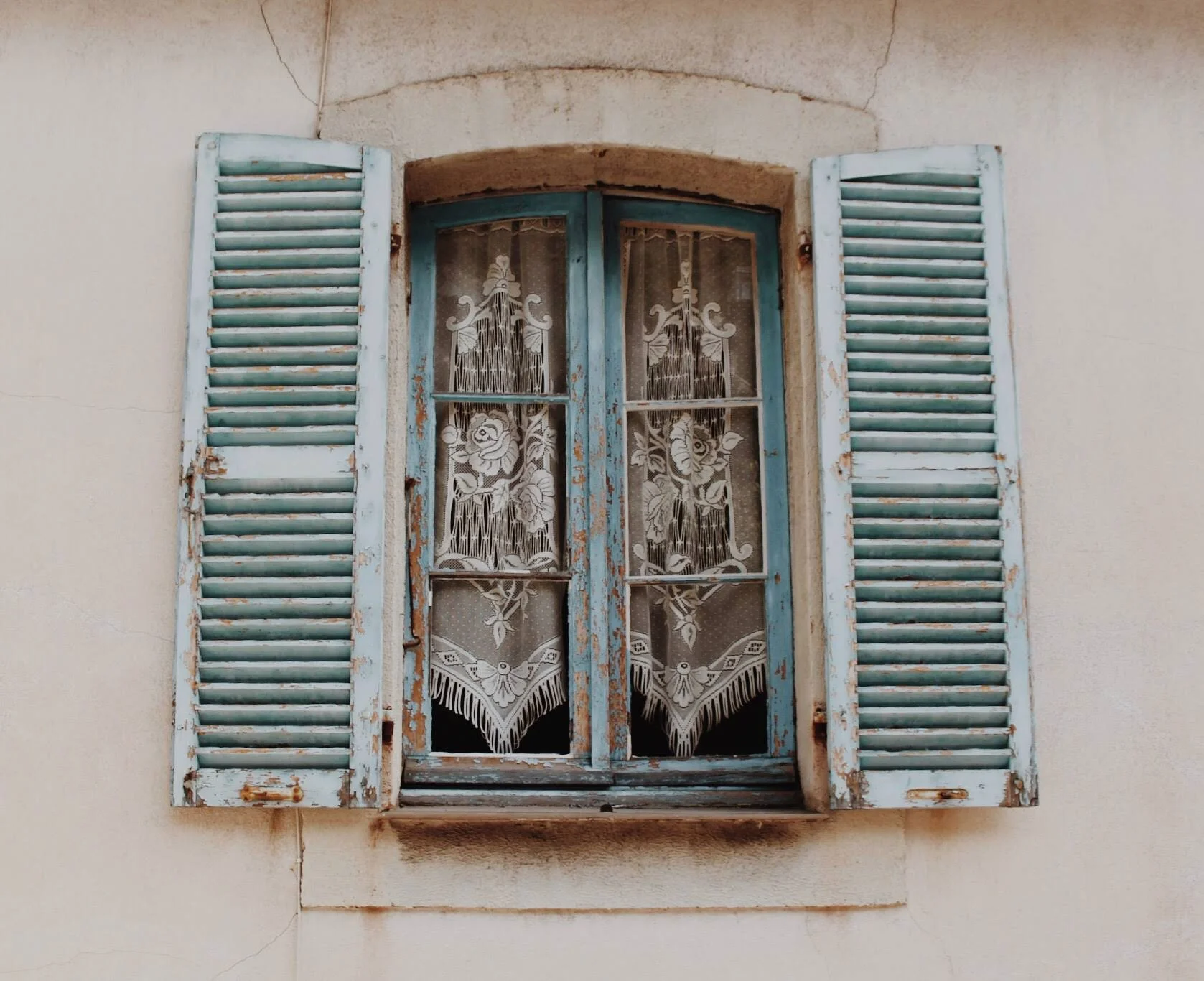 A rustic window with peeling blue paint and wooden shutters flanking both sides. The window panes are adorned with intricate white lace curtains