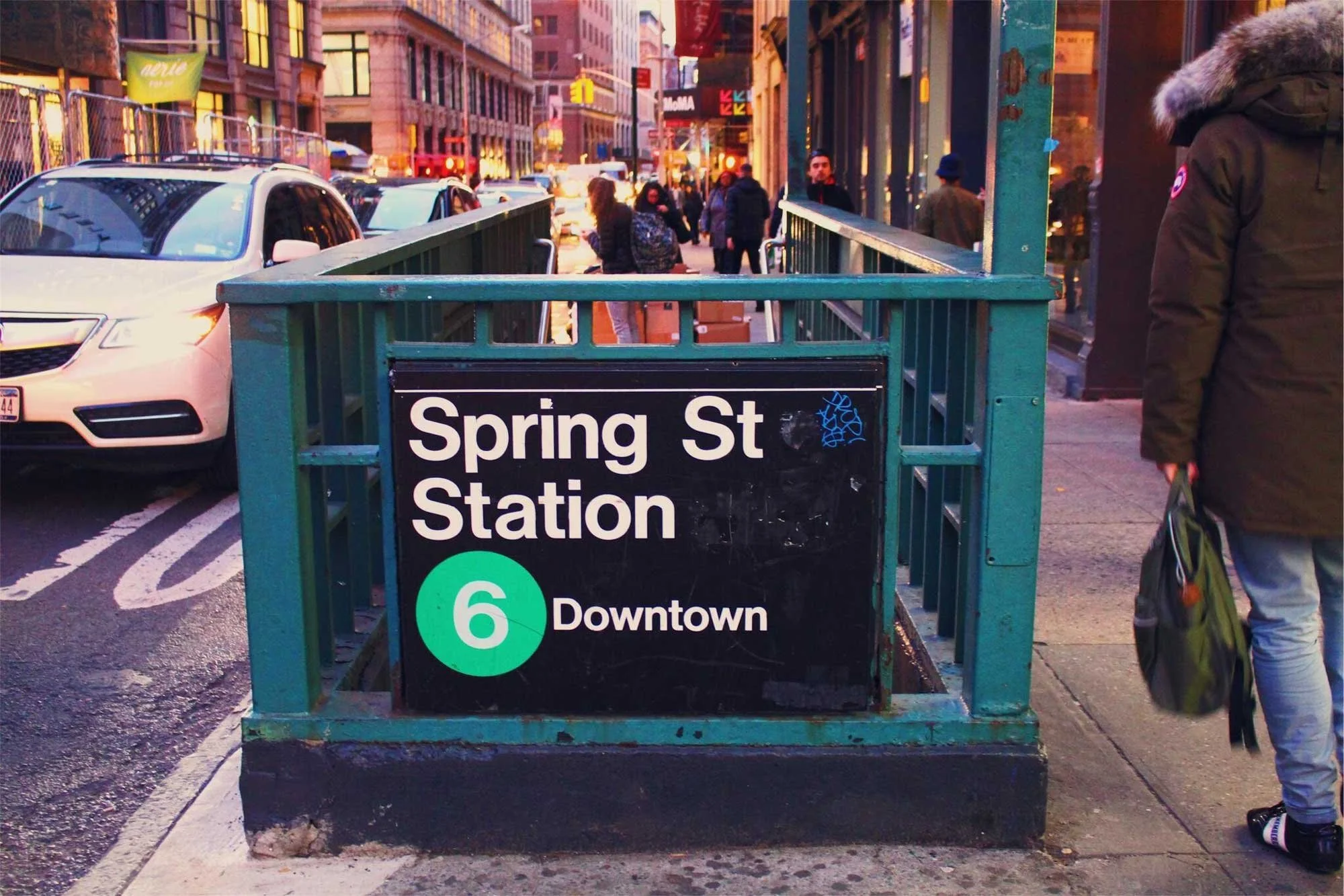 A subway entrance for Spring St Station. Pedestrians and cars are visible on the street, and buildings line the background.