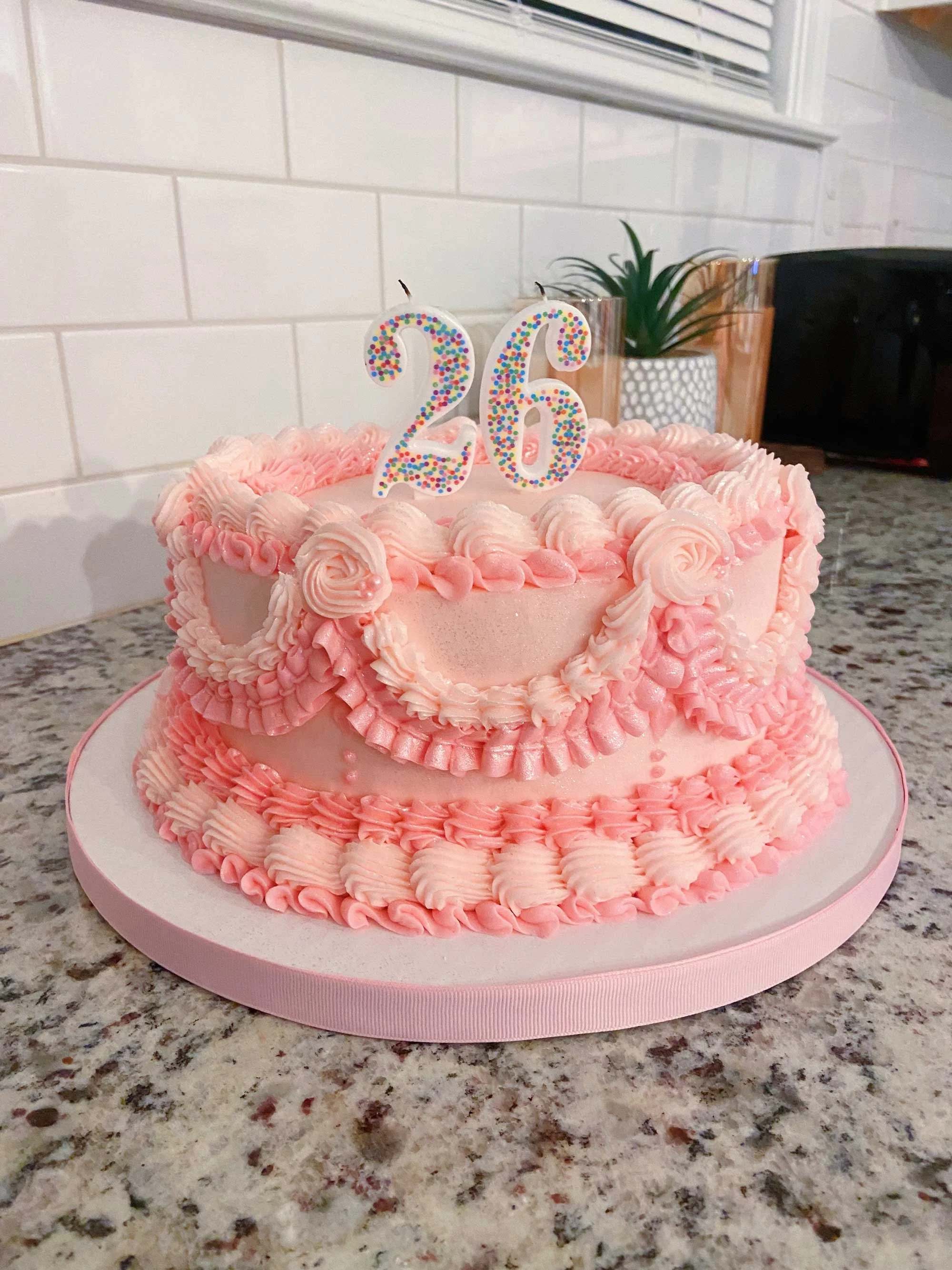 A pink frosted birthday cake adorned with decorative piping sits on a kitchen countertop, topped with number candles reading "26"