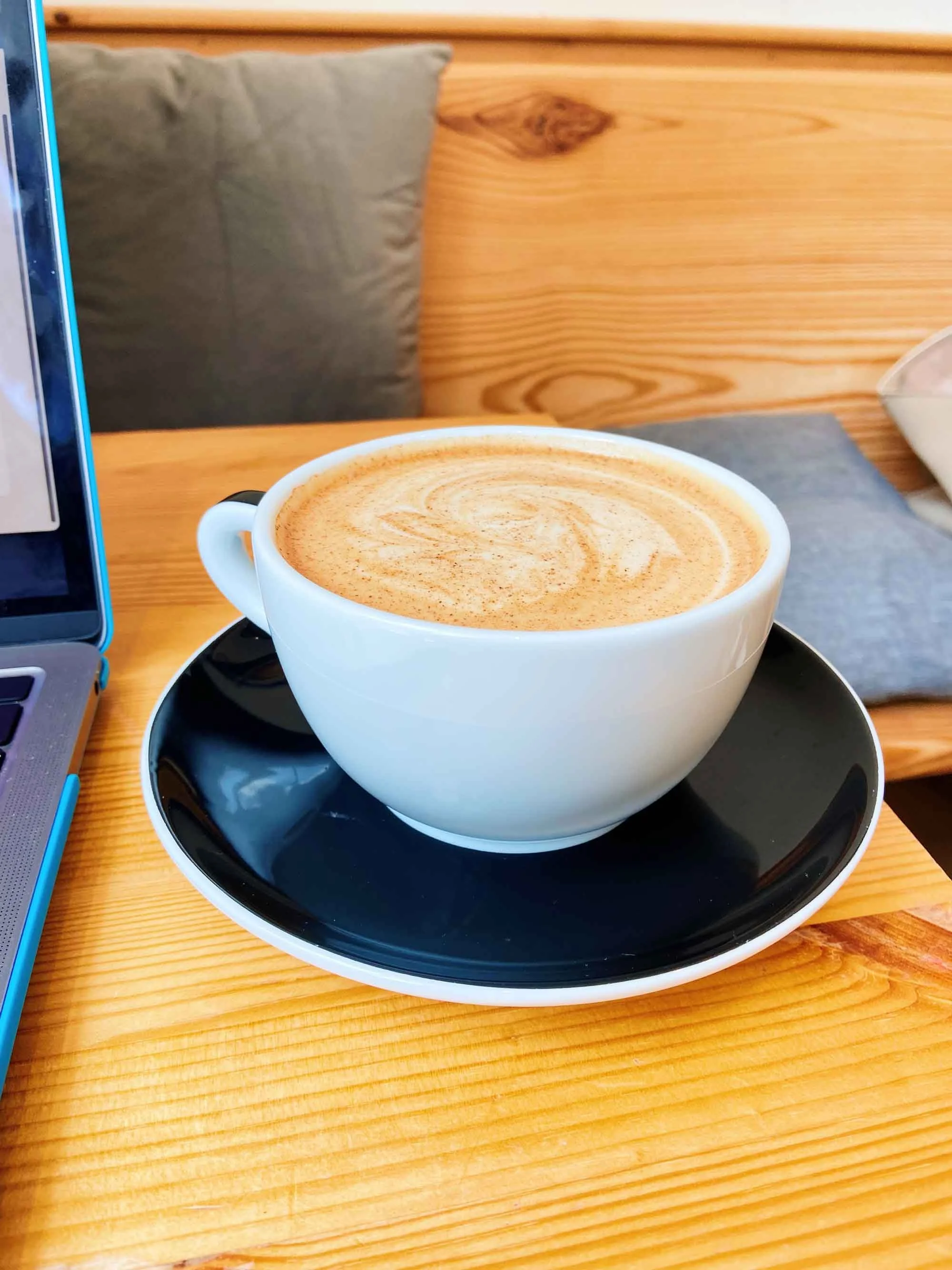 Cup of coffee with frothy milk art sits on a black saucer on a wooden table. Part of a laptop is visible on the left side