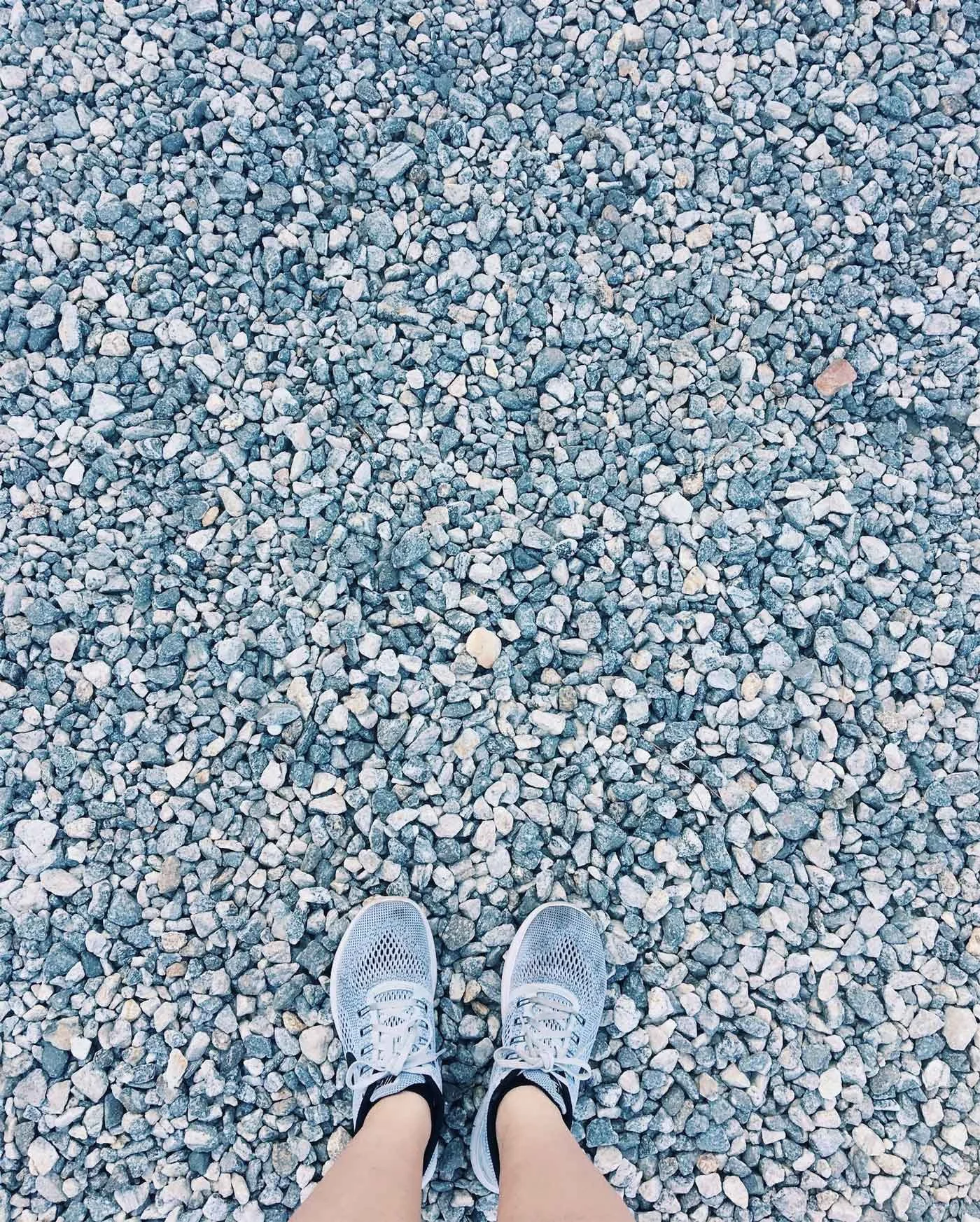 A person wearing light gray sneakers stands on a gravel surface
