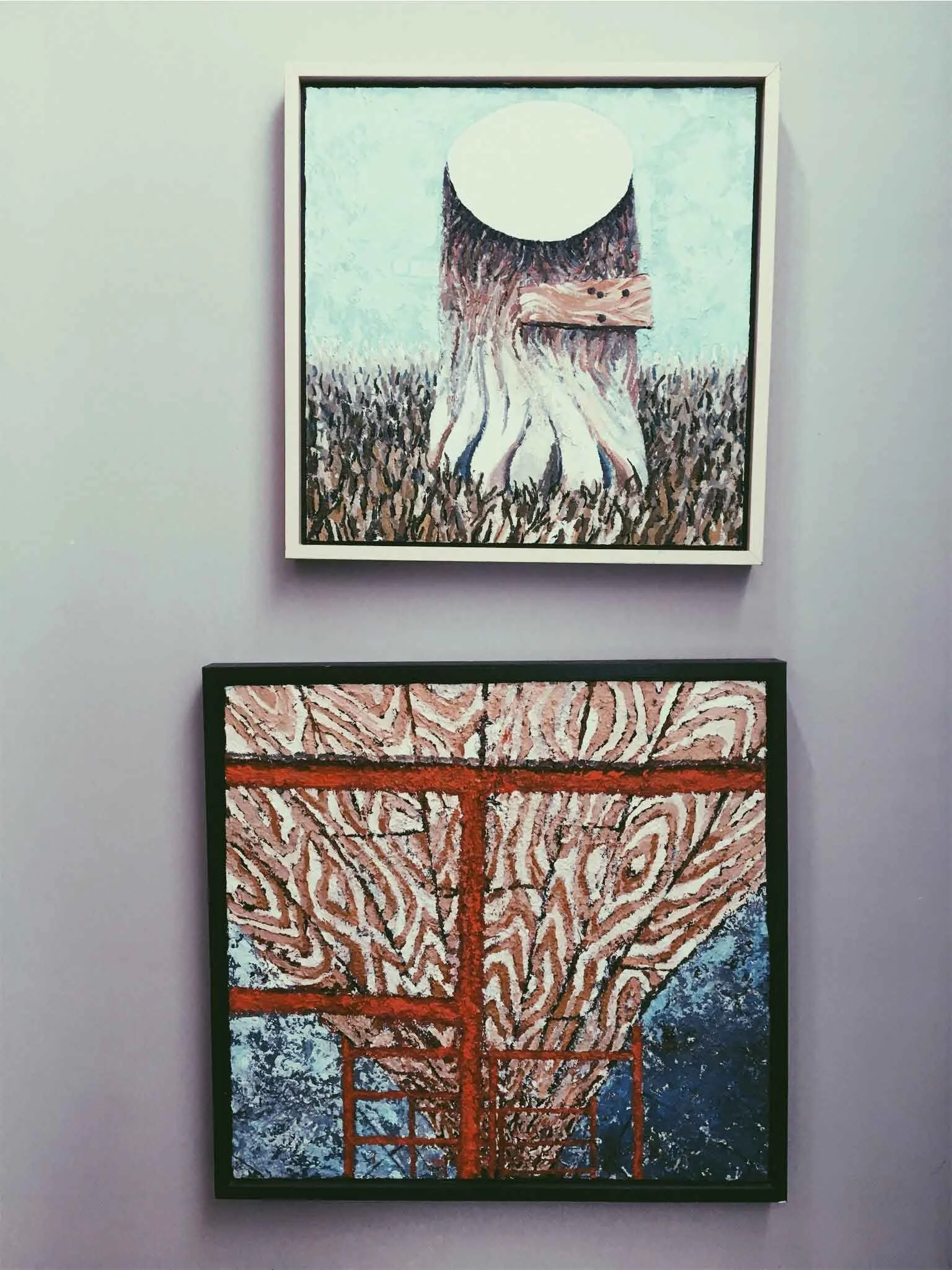 Two framed paintings hang on a wall. The top painting features a tree stump standing amidst grass. The bottom painting includes intertwined red and white lines, resembling wooden planks.