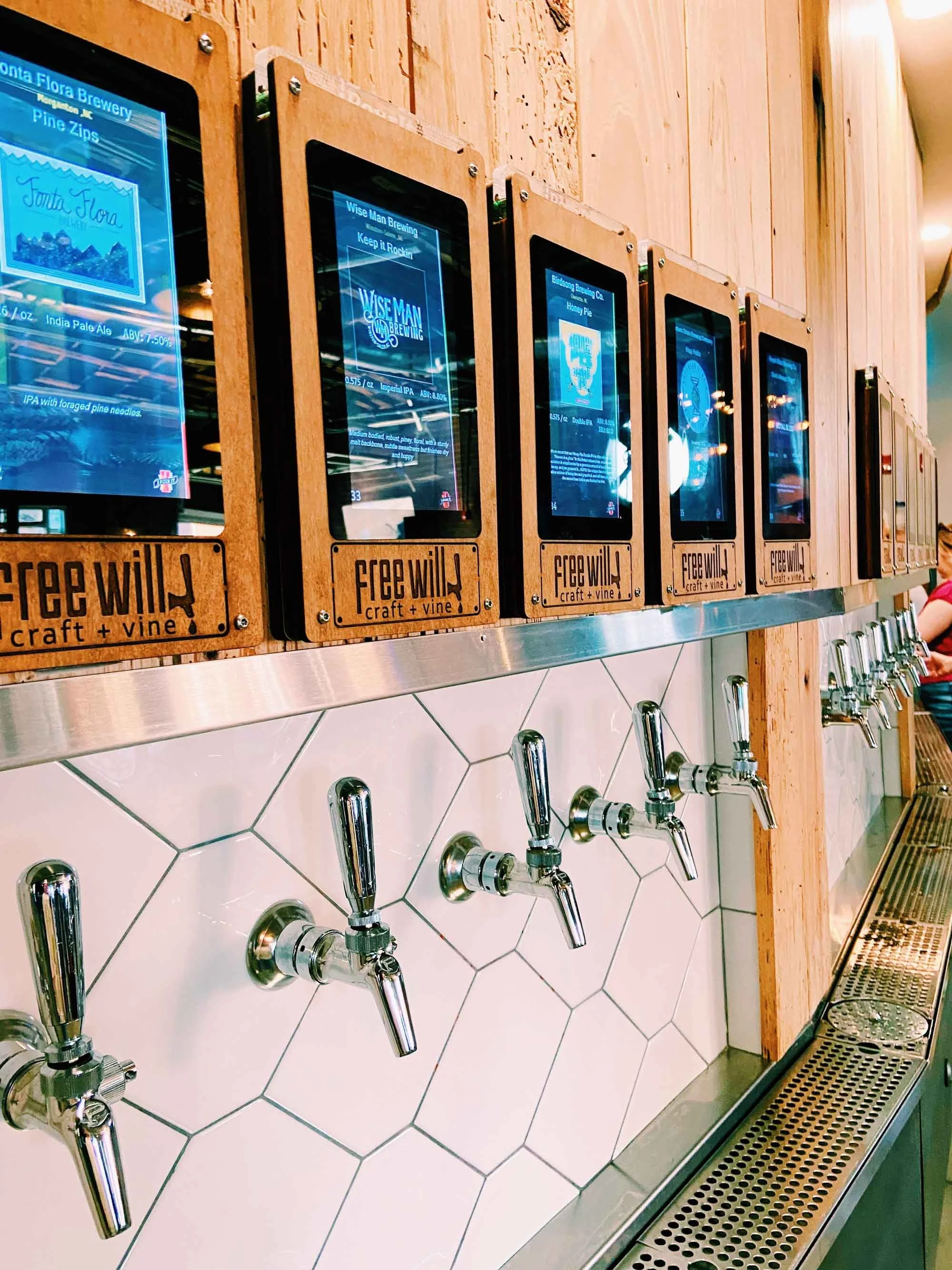 Row of self-service beer taps mounted on a wall with digital screens above each tap, displaying different beer options