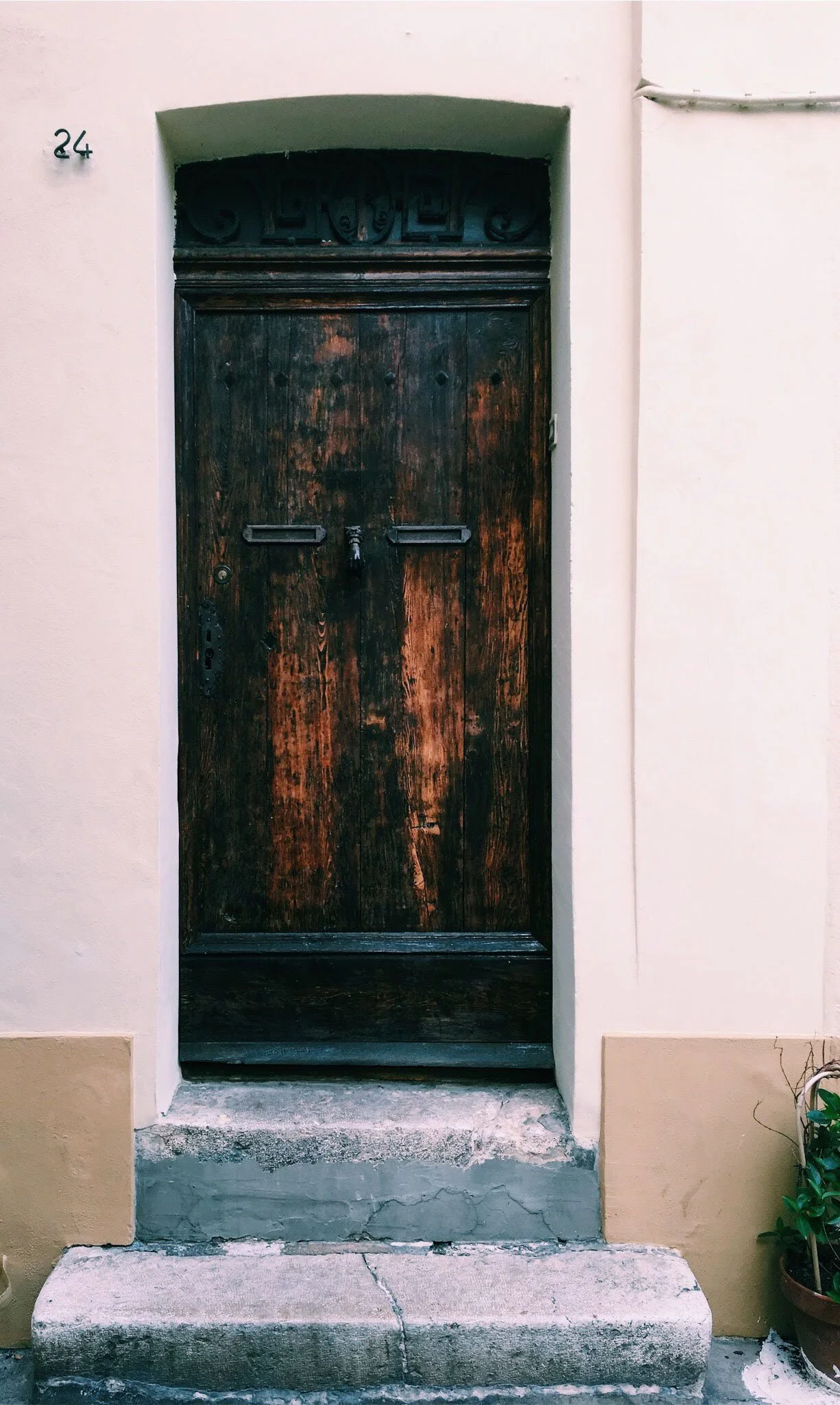 A weathered, dark wooden door set in a pale stucco wall with the number 24 to the left of the doorframe