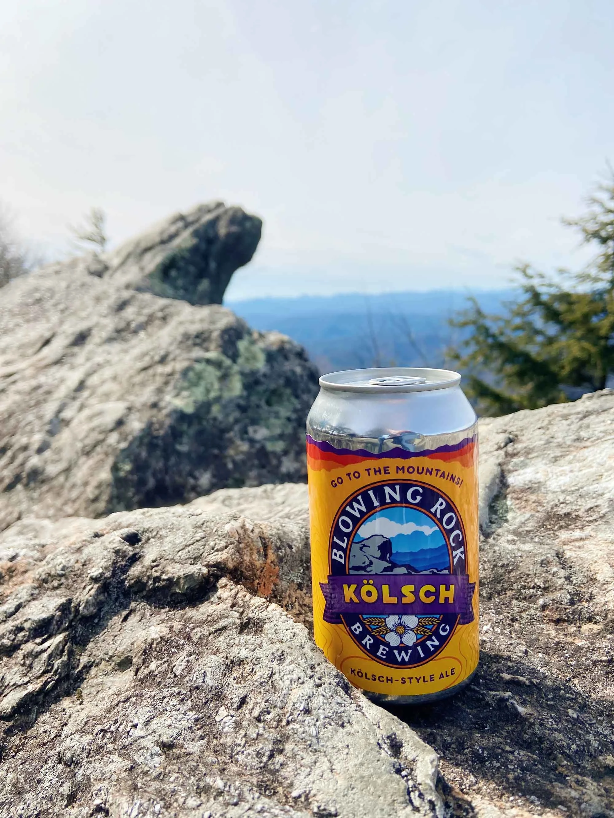 A can of Blowing Rock Kölsch-Style Ale rests on a large rock with a scenic mountain range and blue sky in the background
