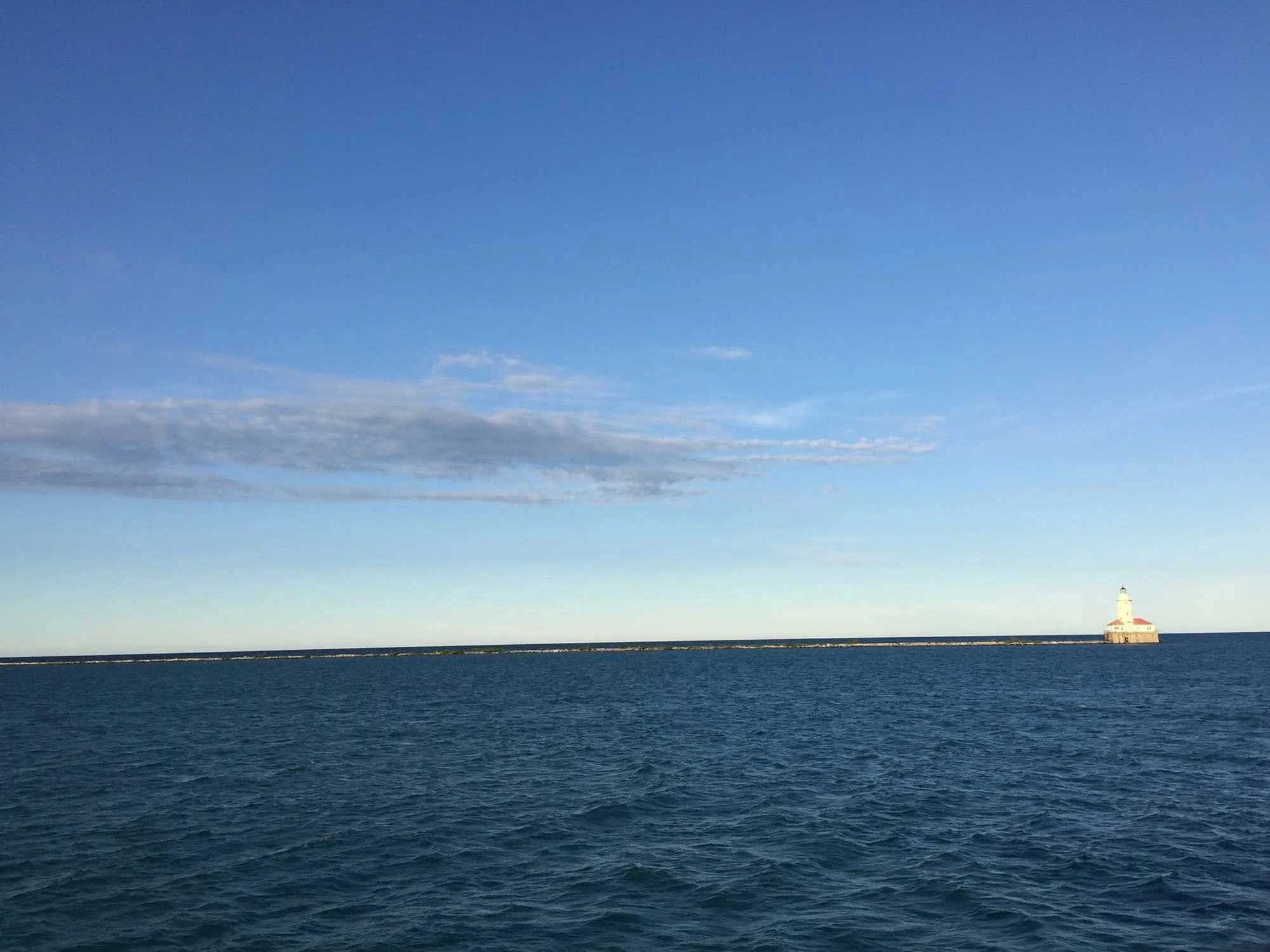 A vast, calm sea under a blue sky. On the horizon, there is a small, distant lighthouse