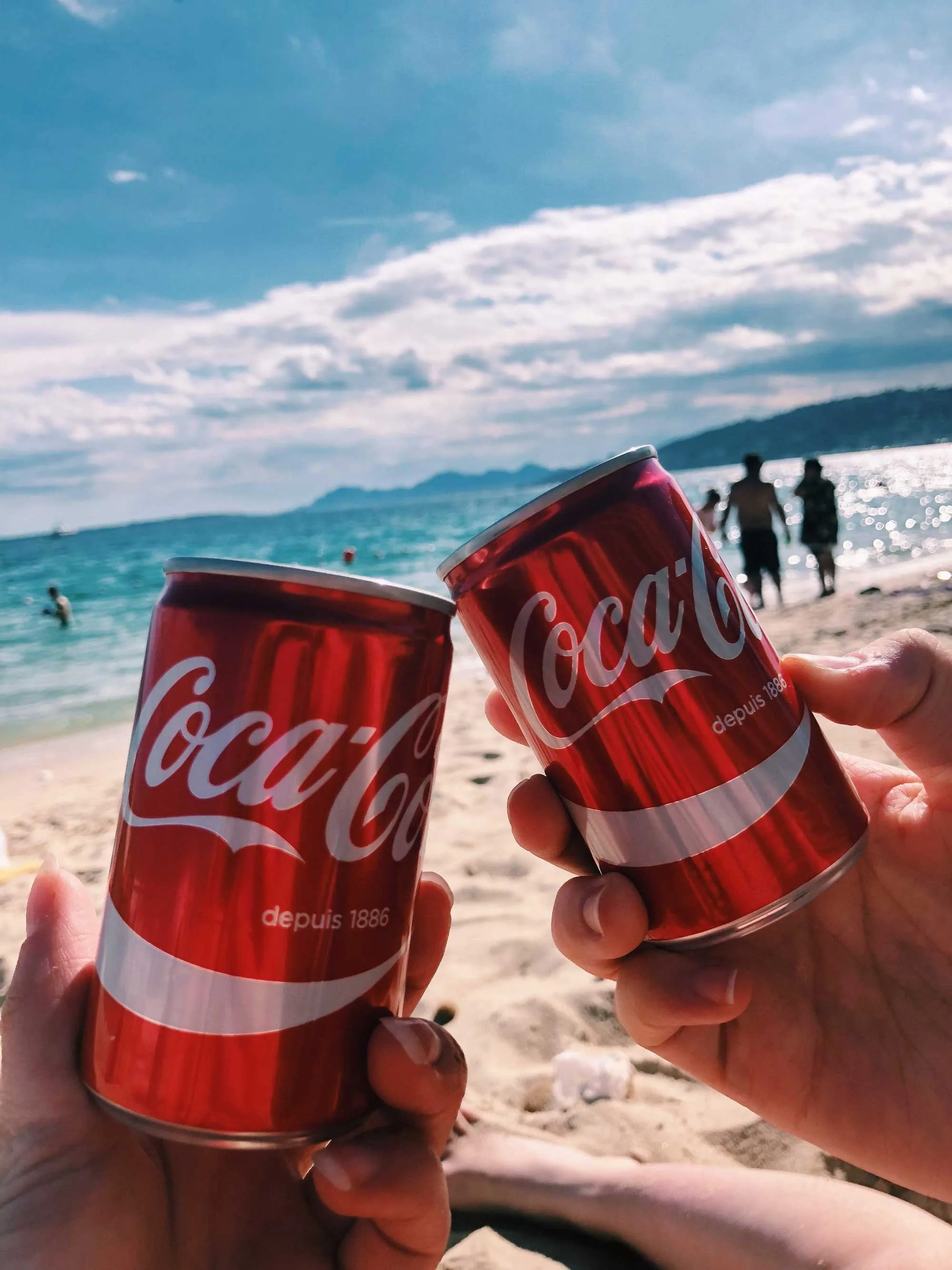 Two hands holding mini red Coca-Cola cans with the ocean and beach in the background on a sunny, partly cloudy day. 