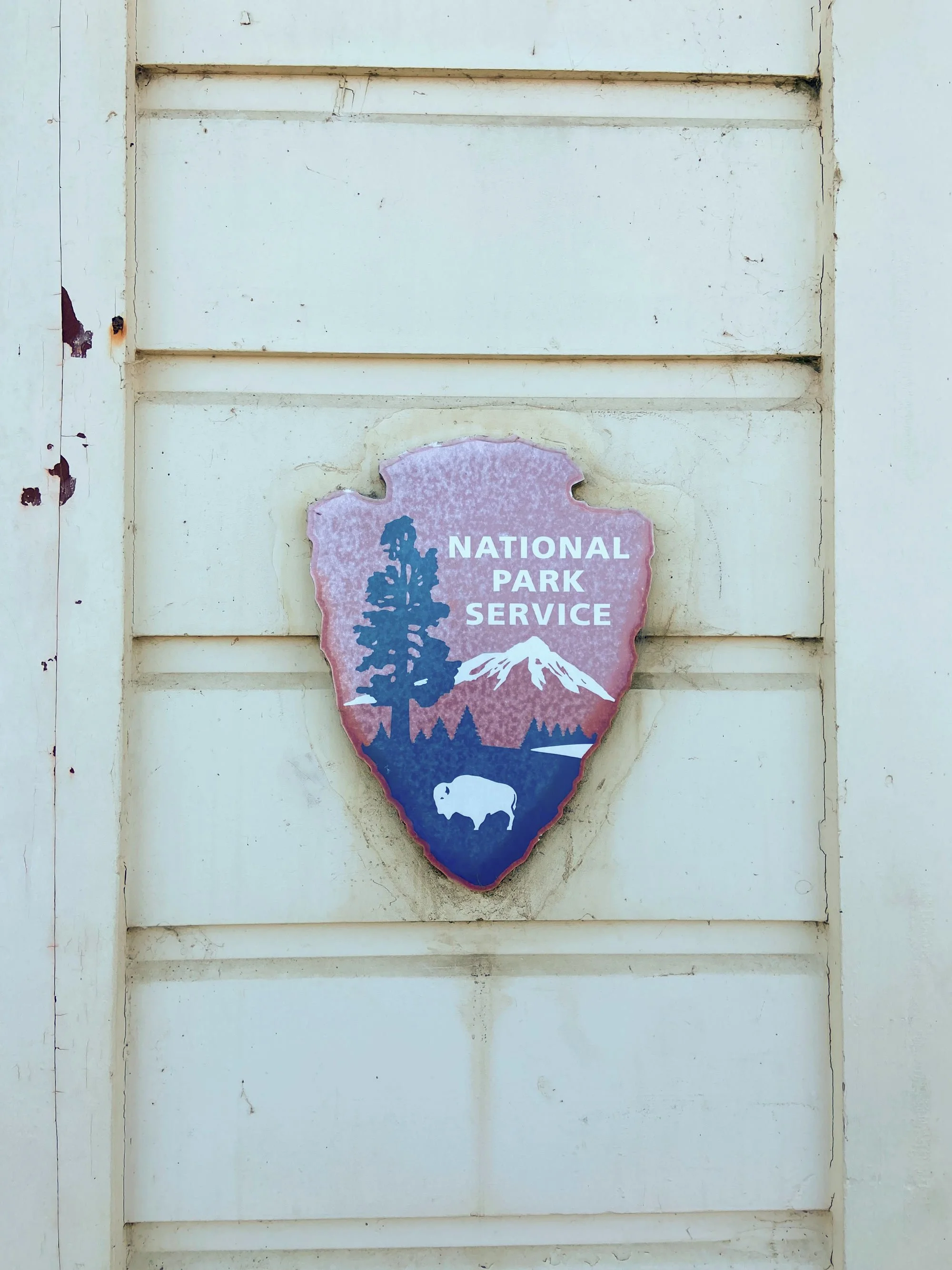 Weathered sign mounted on a white wooden wall. The sign reads "National Park Service" and depicts a tree, mountains, and a bison