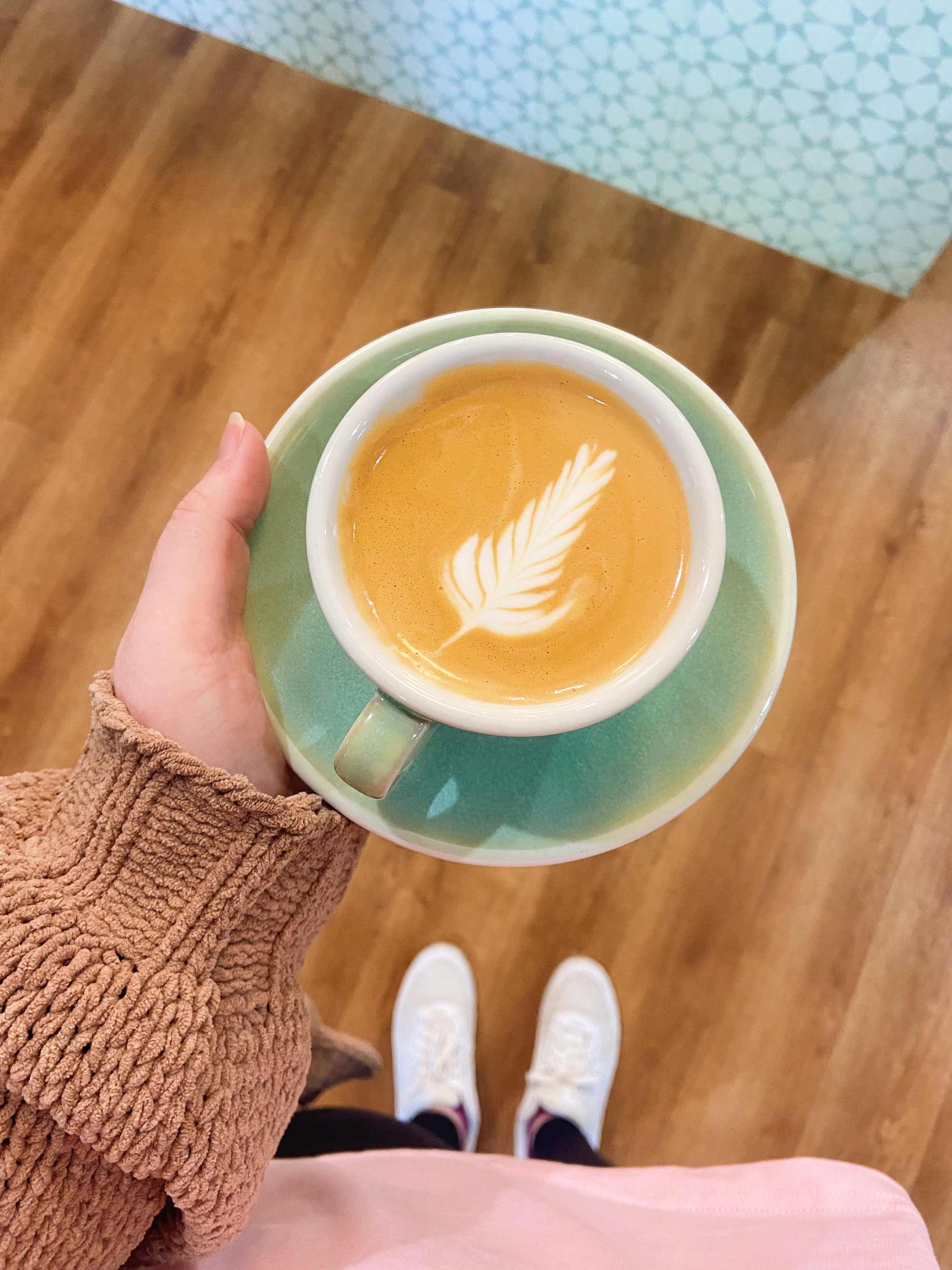 Person wearing a brown knit sweater and white sneakers holding a green saucer and cup filled with a latte, featuring a fern latte art design on top