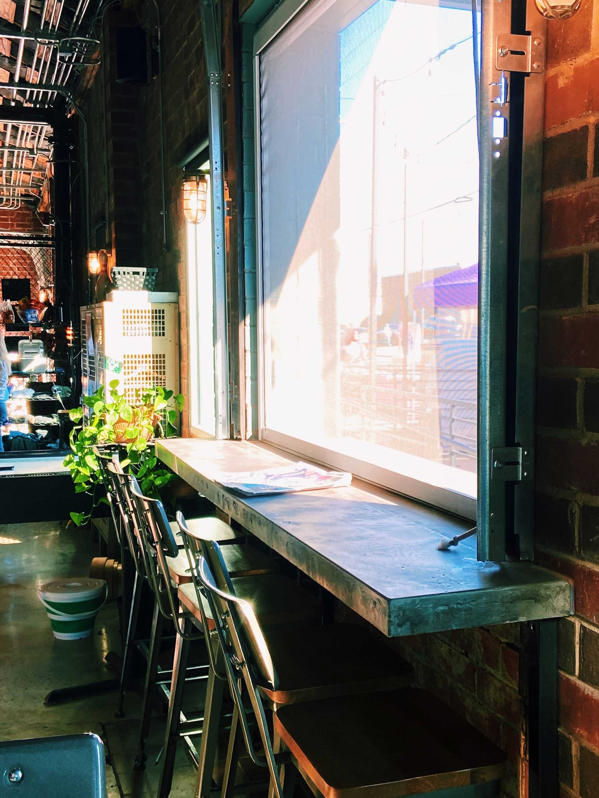 Sunlit indoor cafe scene with high window. Decor is industrial with brick walls and metal accents.