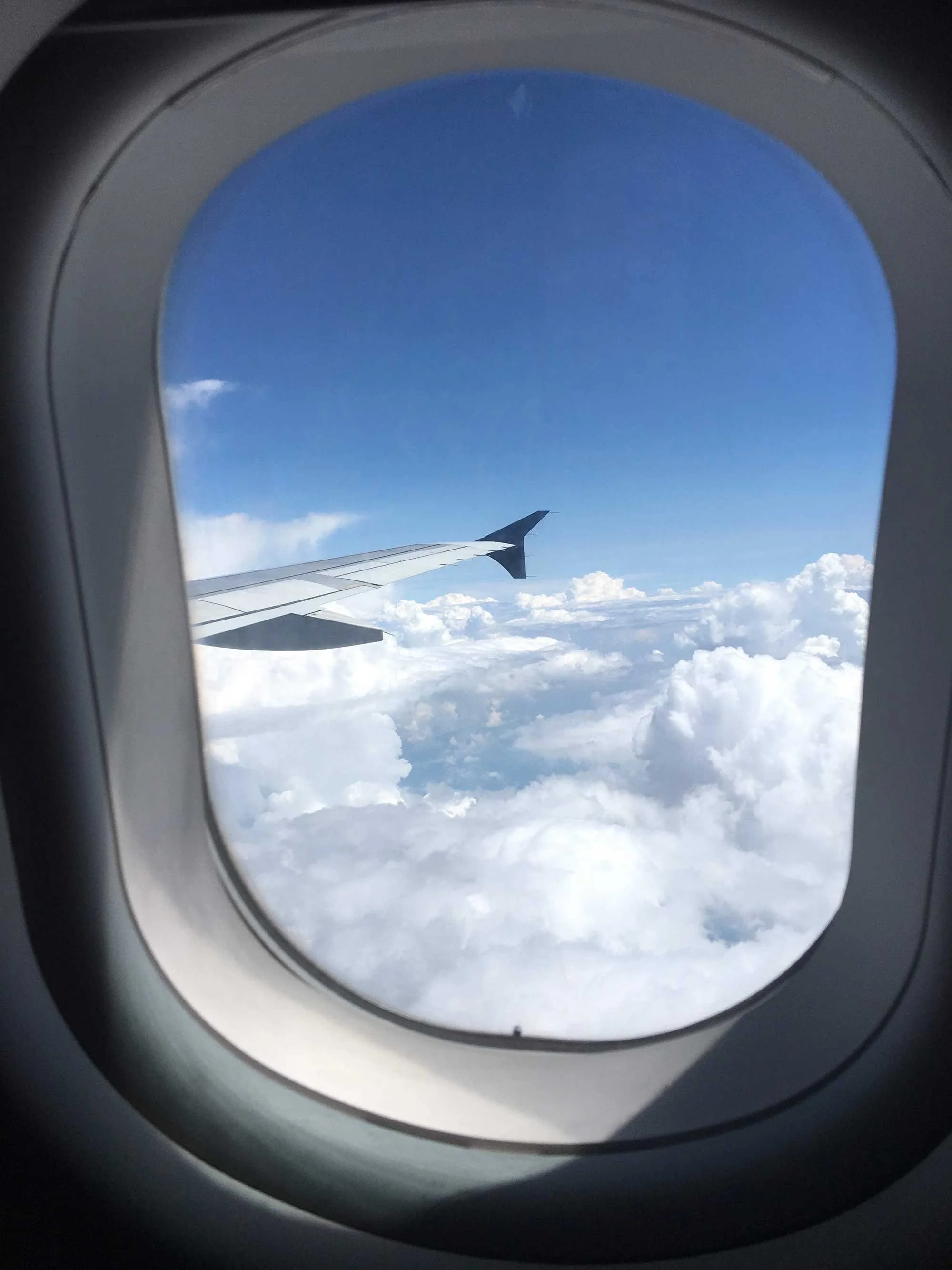 View through an airplane window showing an airplane wing against a backdrop of bright blue sky and fluffy white clouds