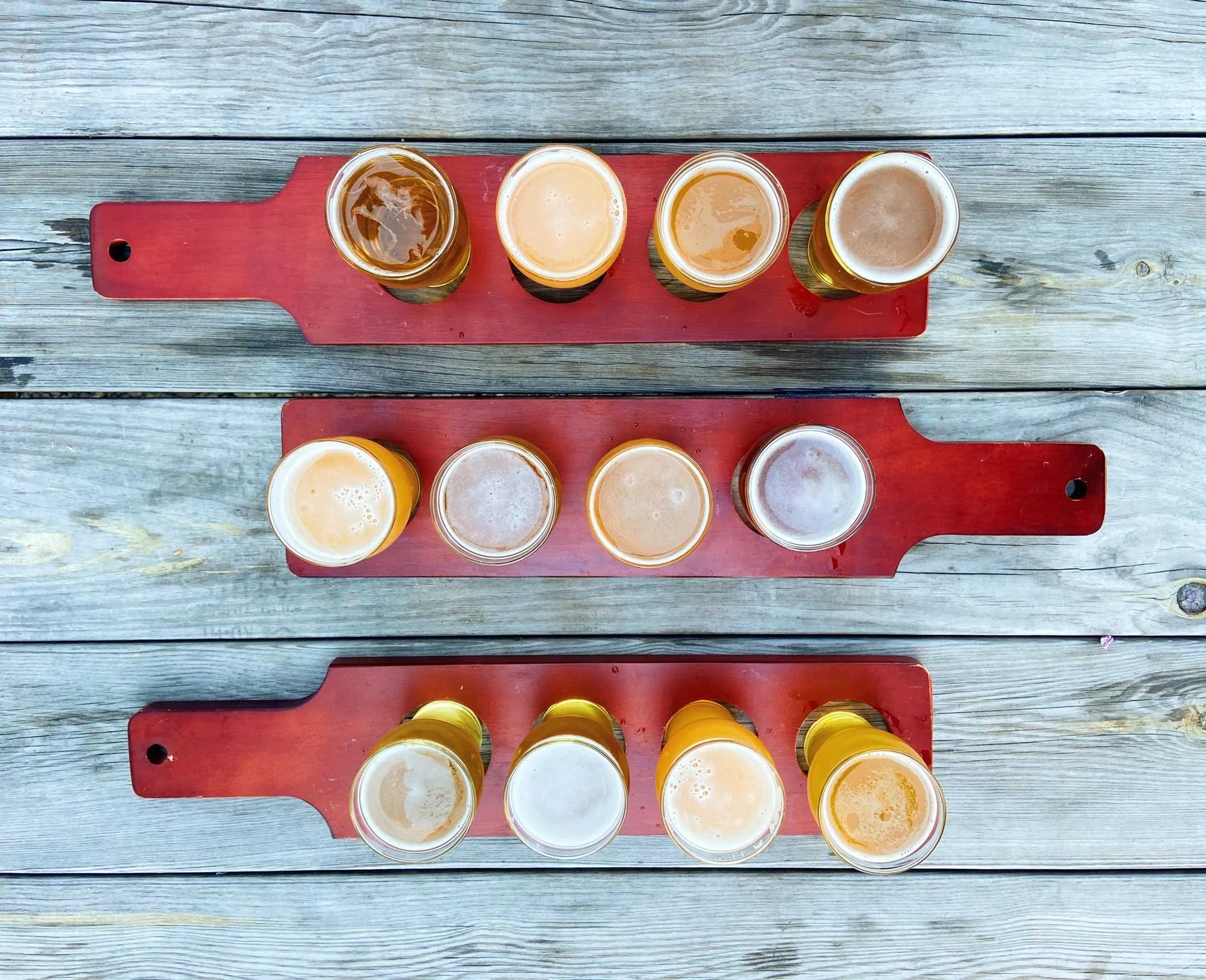 Three wooden flight boards, each holding four glasses of different types of beer, are arranged on a rustic wooden table