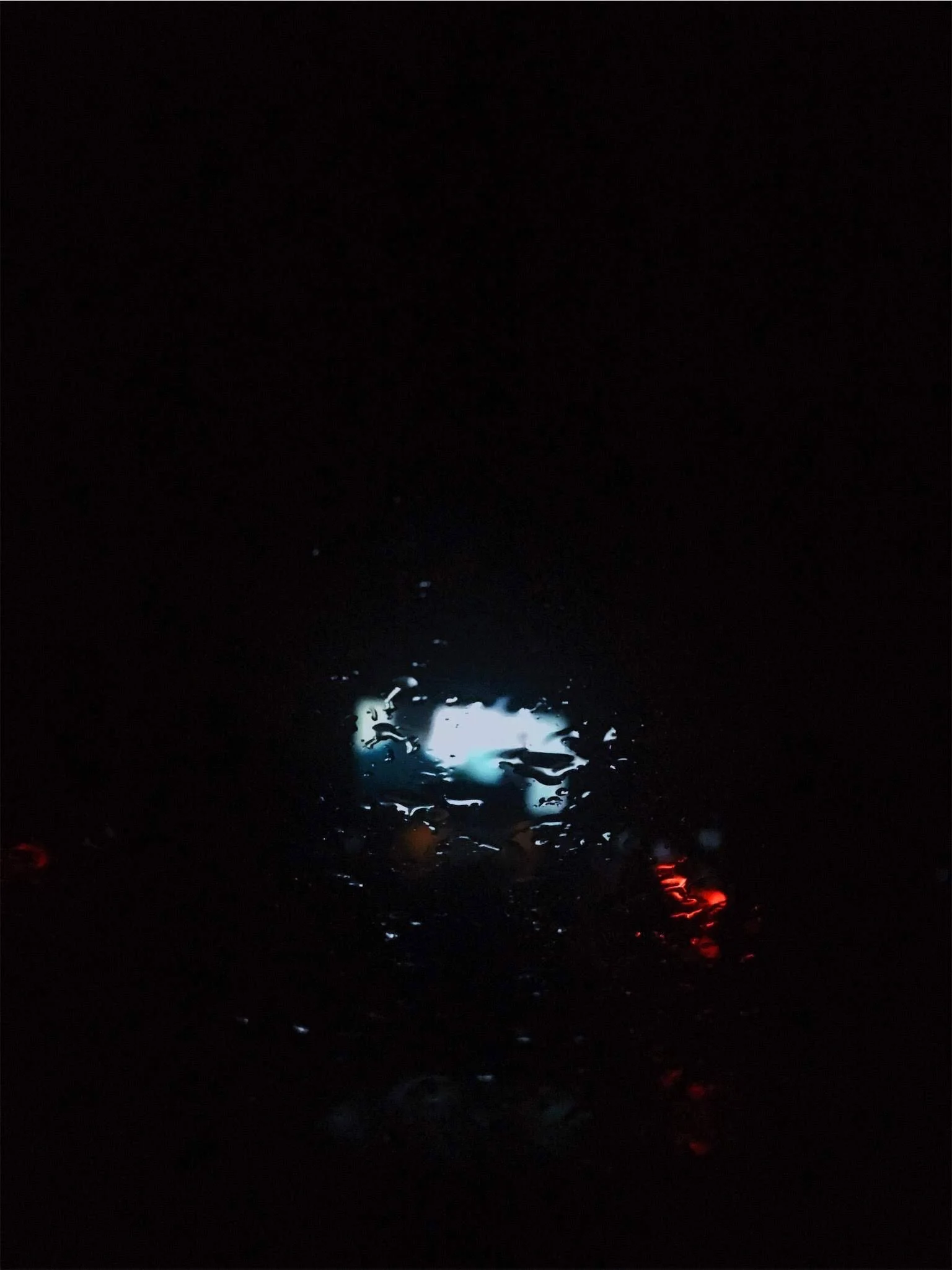 A rain-covered window at night with small, scattered red and amber lights appearing through the raindrops, contributing to a moody atmosphere