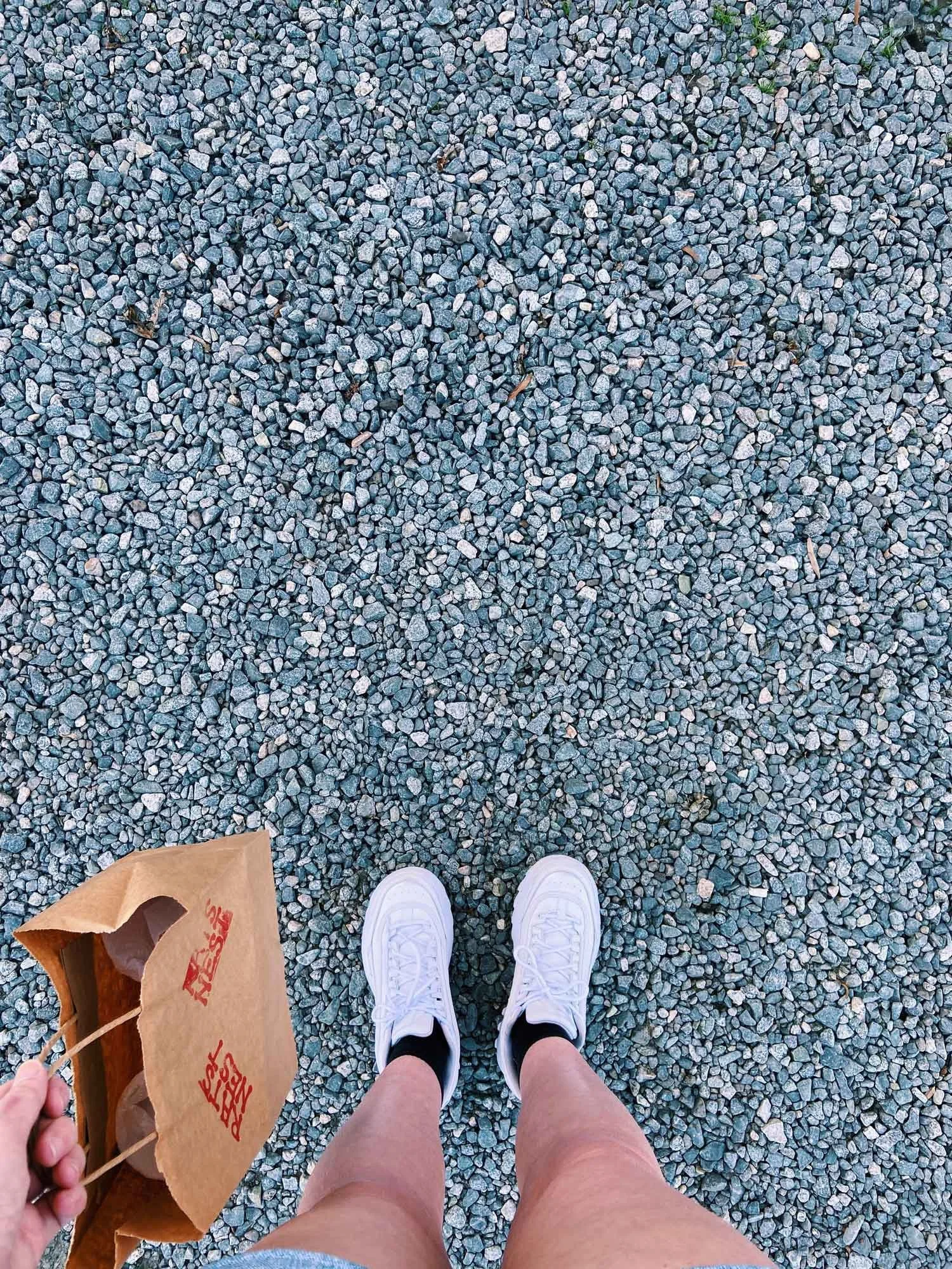Person stands on gravel, visible from knees down. They wear white sneakers and hold a brown paper bag labeled "RATS NEST"