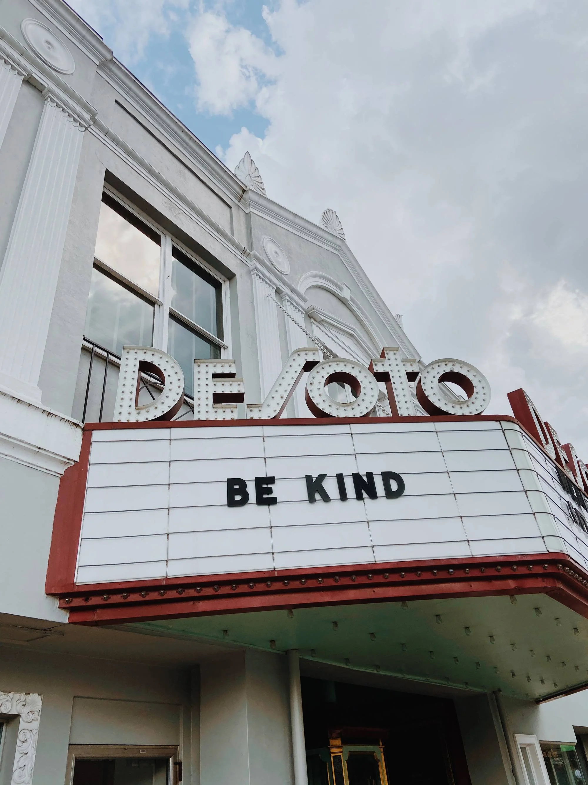 Vintage theater marquee on white building displays message "BE KIND" beneath theater name "DESOTO"