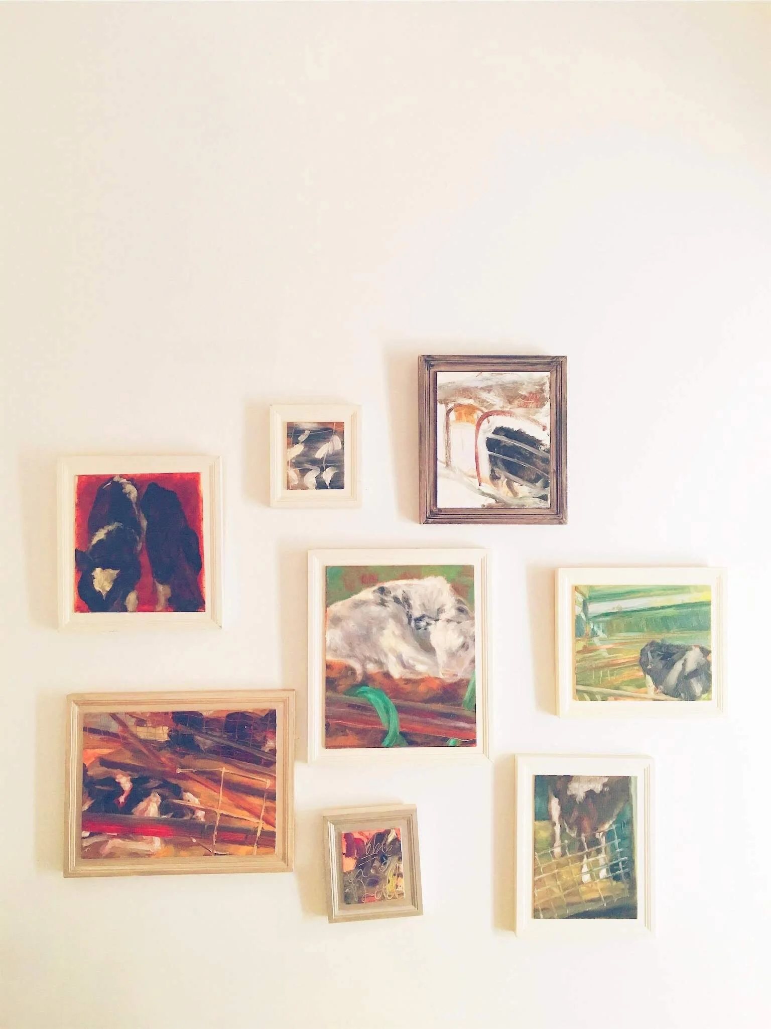 A photo of a light-colored wall displaying various framed paintings depicting abstract and impressionistic scenes, including animals and landscapes