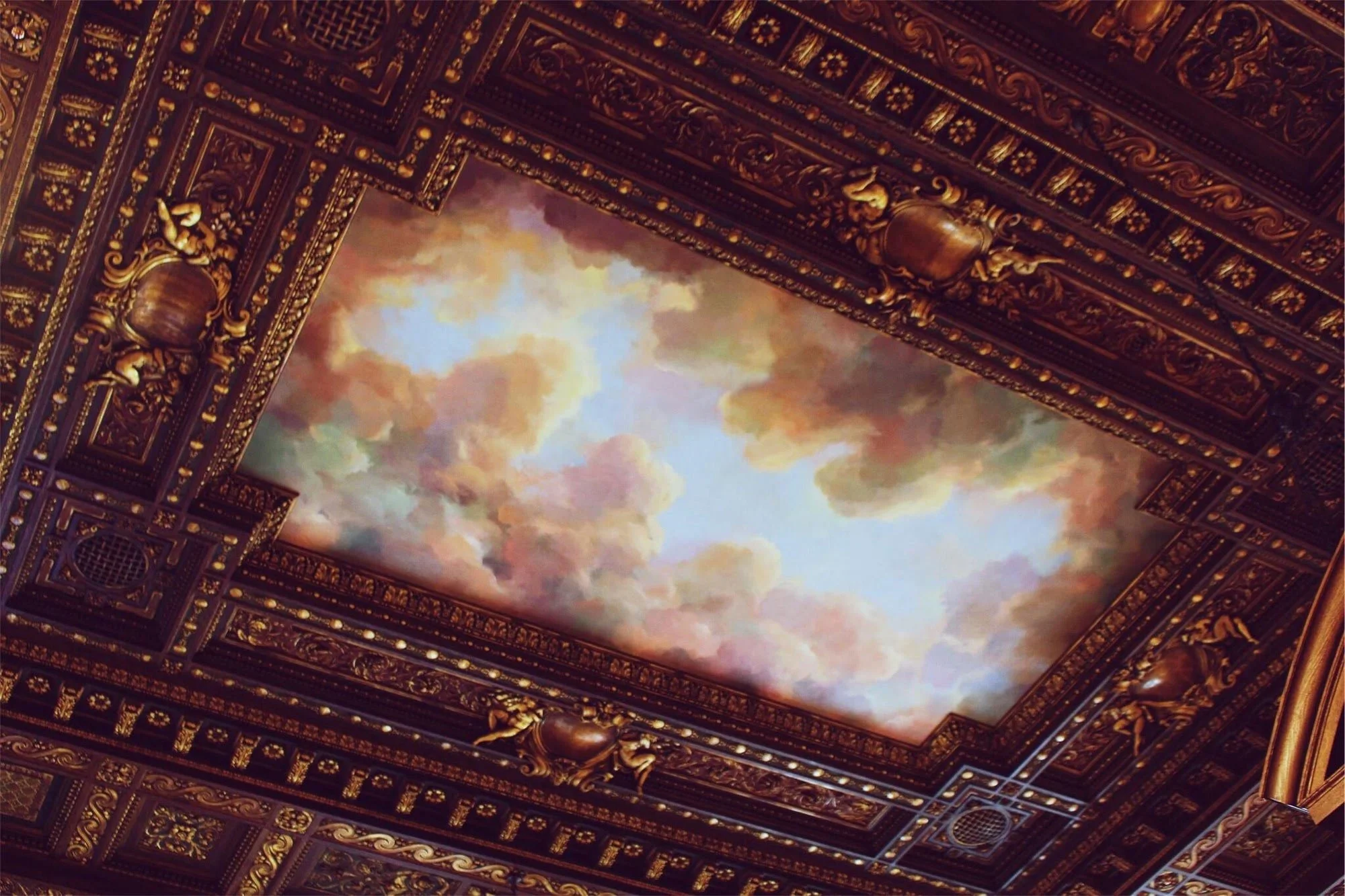 Richly decorated ceiling with intricate gold details surrounds a painting depicting colorful, softly blended clouds against a bright sky
