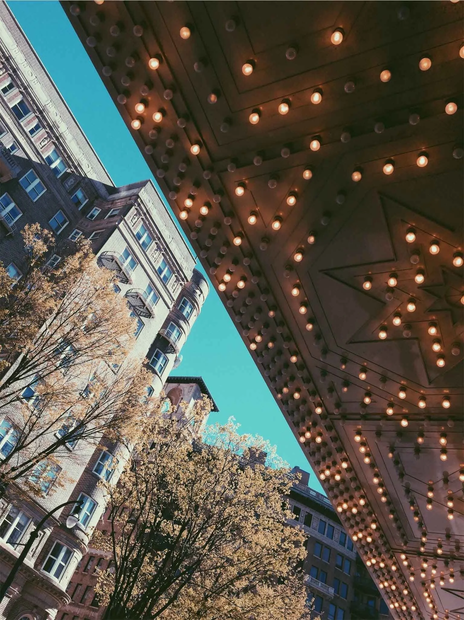 A city street view looking upward, capturing the underside of a theater marquee adorned with lit bulbs arranged in geometric patterns