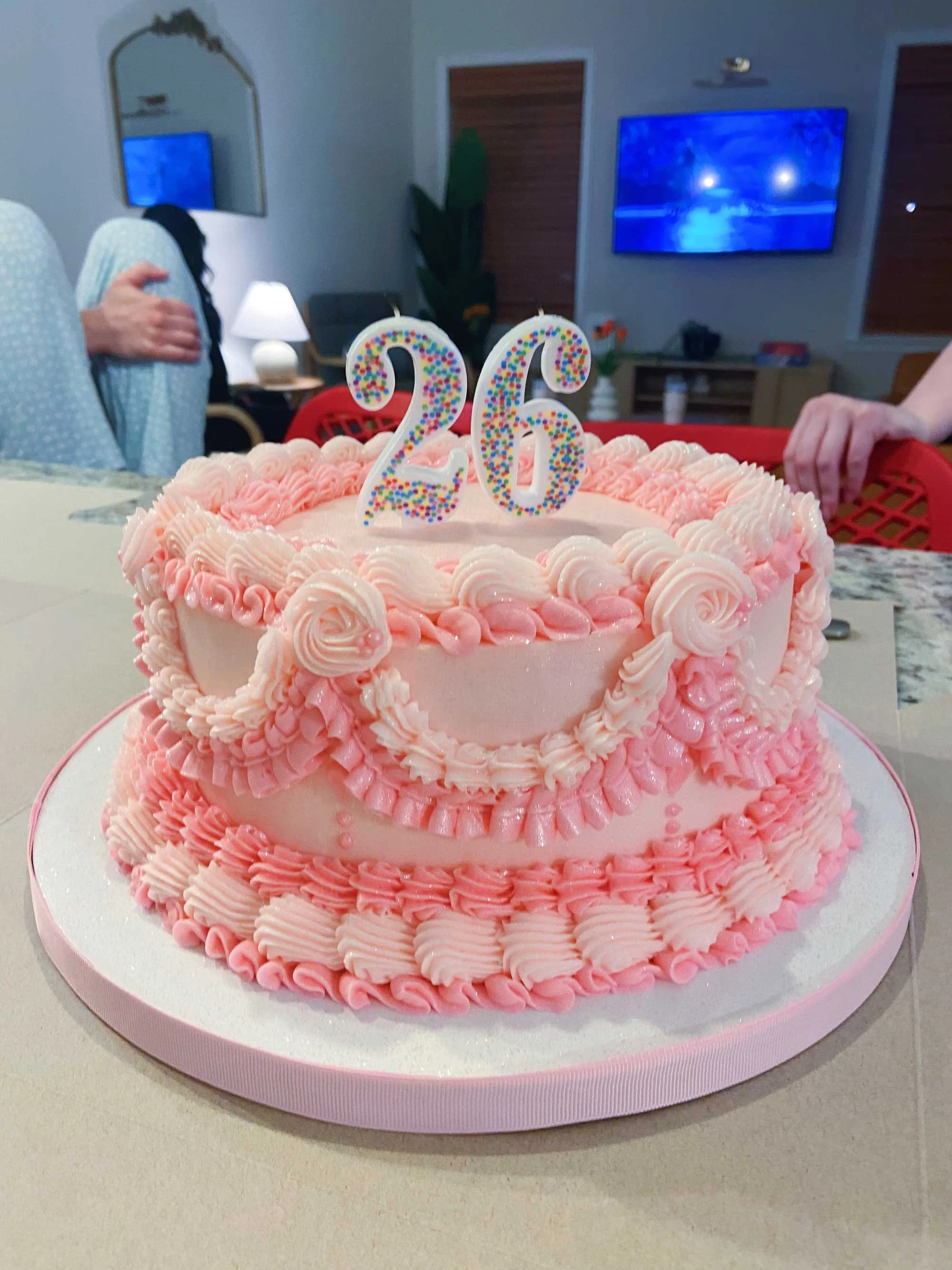 Pink frosted cake with decorative icing, topped with number candles "26" in front of a blurred room setting including a TV screen and furniture