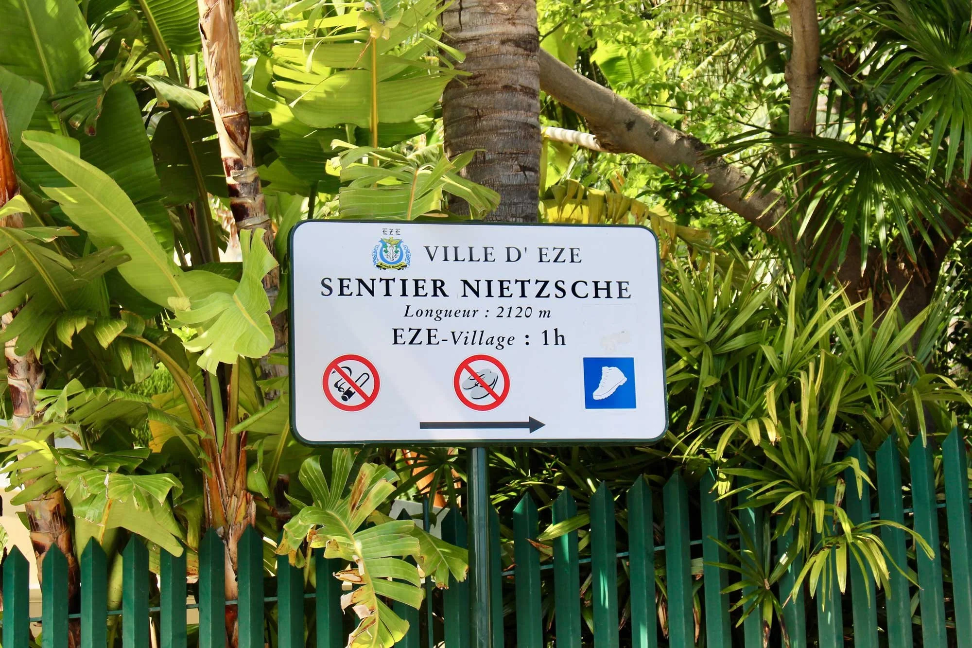A white signboard in front of lush greenery directs to "Sentier Nietzsche" in Èze