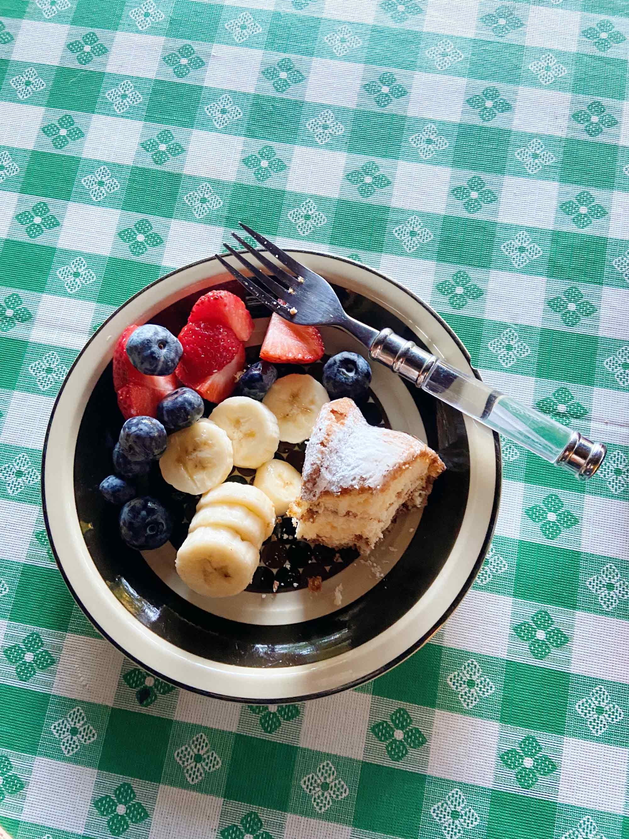 Plate with slices of banana, berries, and a powdered pastry on green and white checkered tablecloth