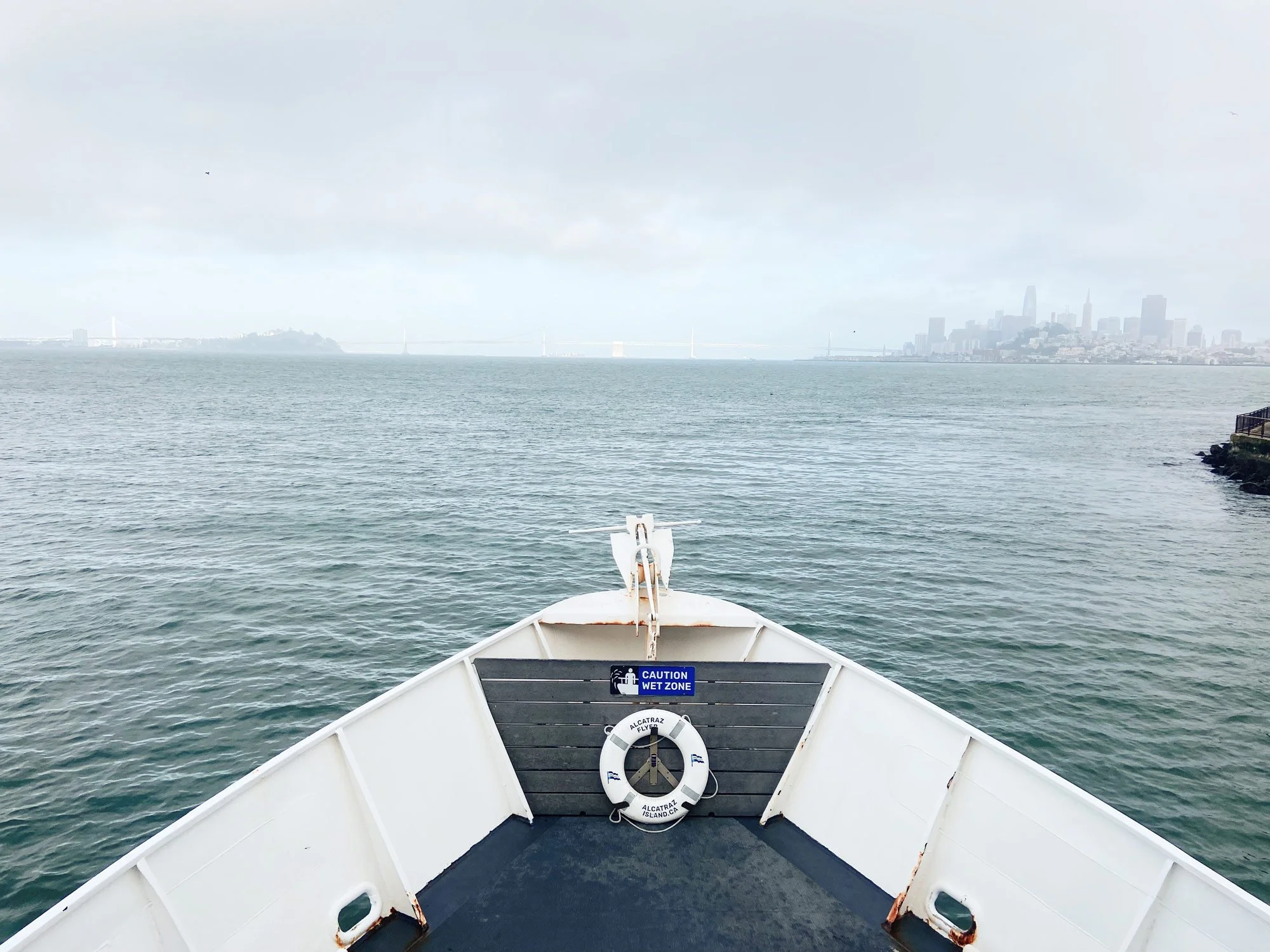 The bow of a boat moves through calm waters with a hazy city skyline in the background