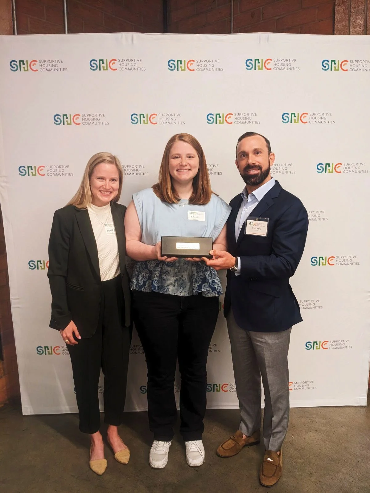 Three people stand in front of a banner that reads "Supportive Housing Communities." Katie Shivers stands in the middle holding a rectangular award, smiling.