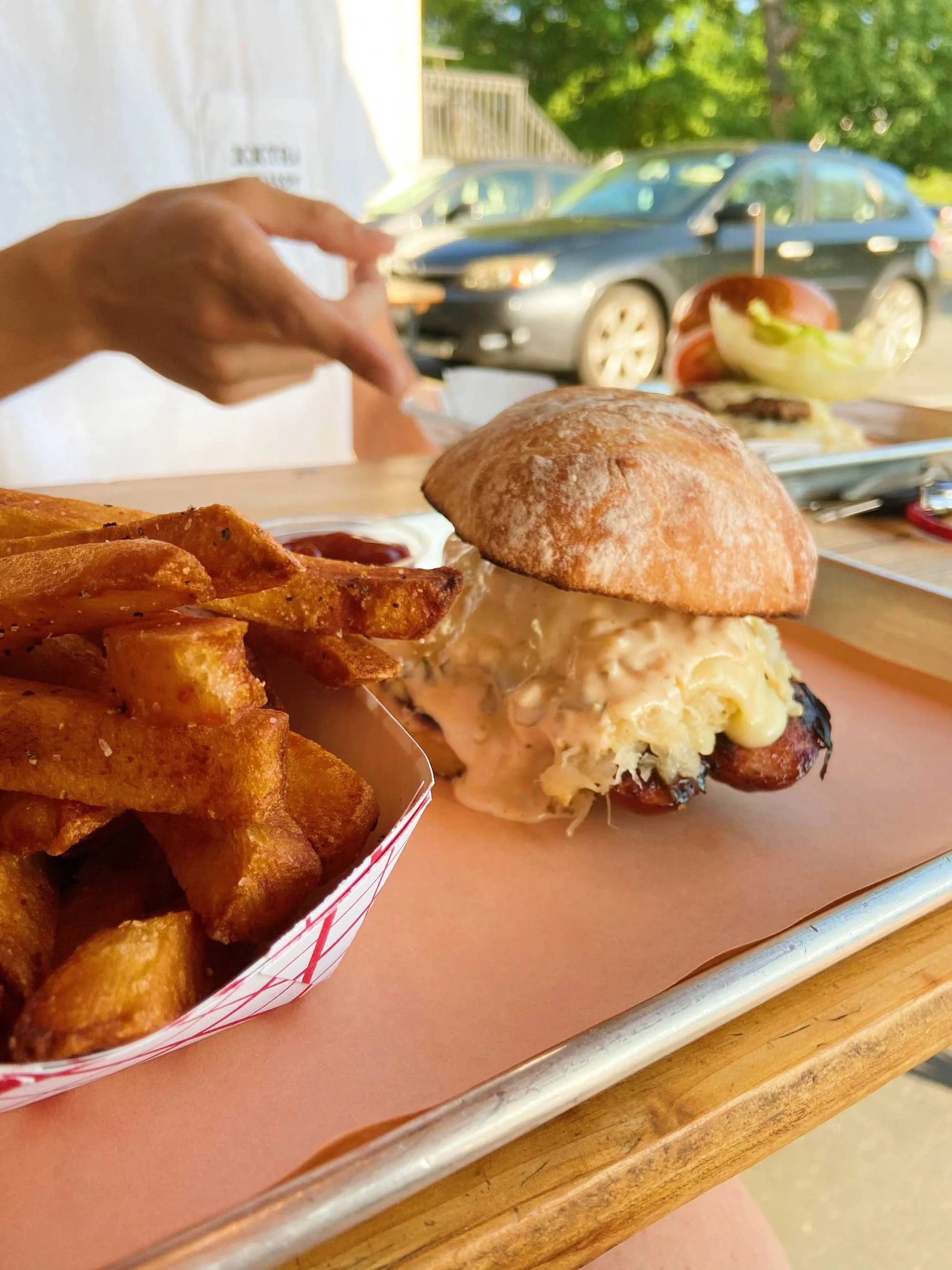 Person points at a tray with a bratwurst sandwich oozing with sauce and filling, paired with a serving of golden-brown fries