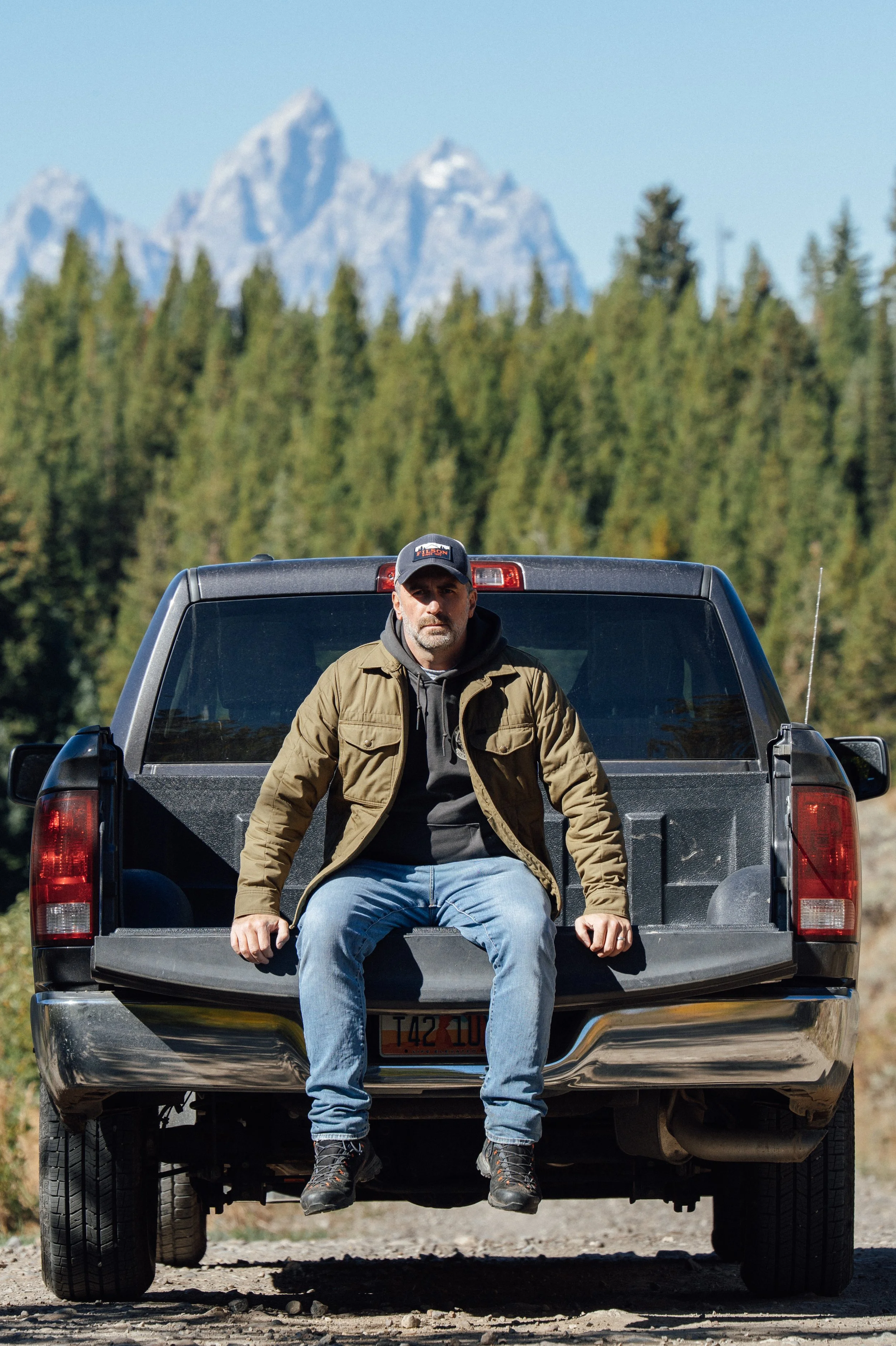 Man in baseball cap sitting in the back of truck with Teton mountains and trees in the distance