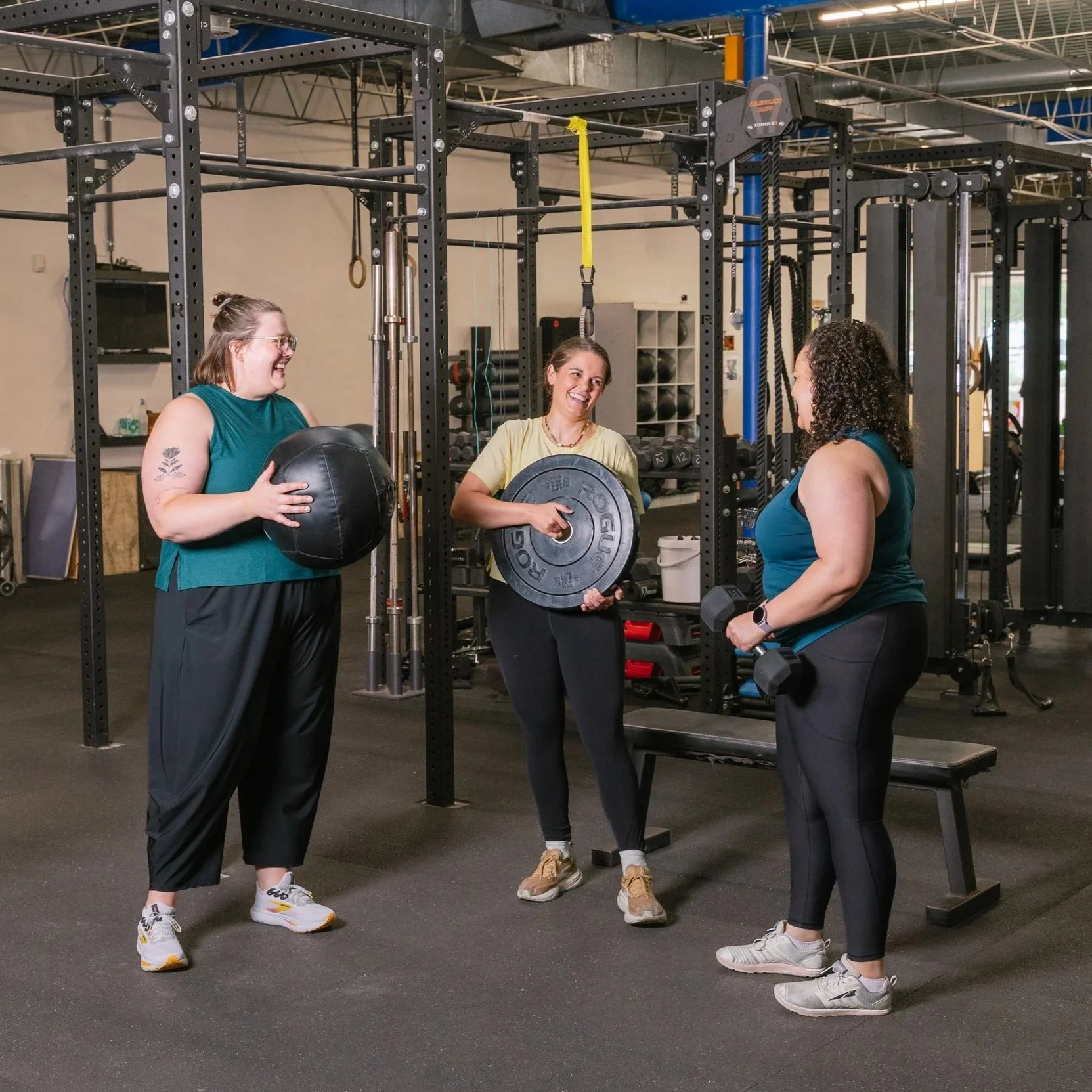 Allison Mosso and two women in the gym, each holding some equipment: a weighted medicine ball, a weight plate, and a dumbbell.