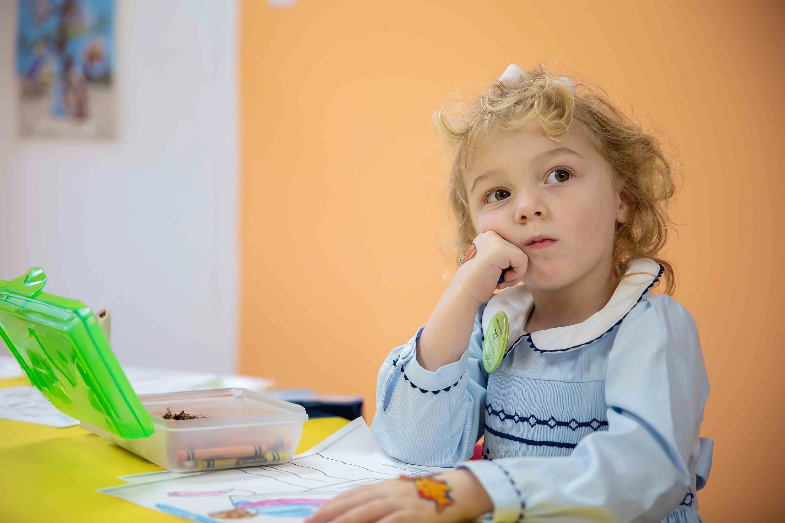 Young girl with glasses and a yellow polka dot dress sitting and listening attentively at a classroom or event, with other children blurred in the background.