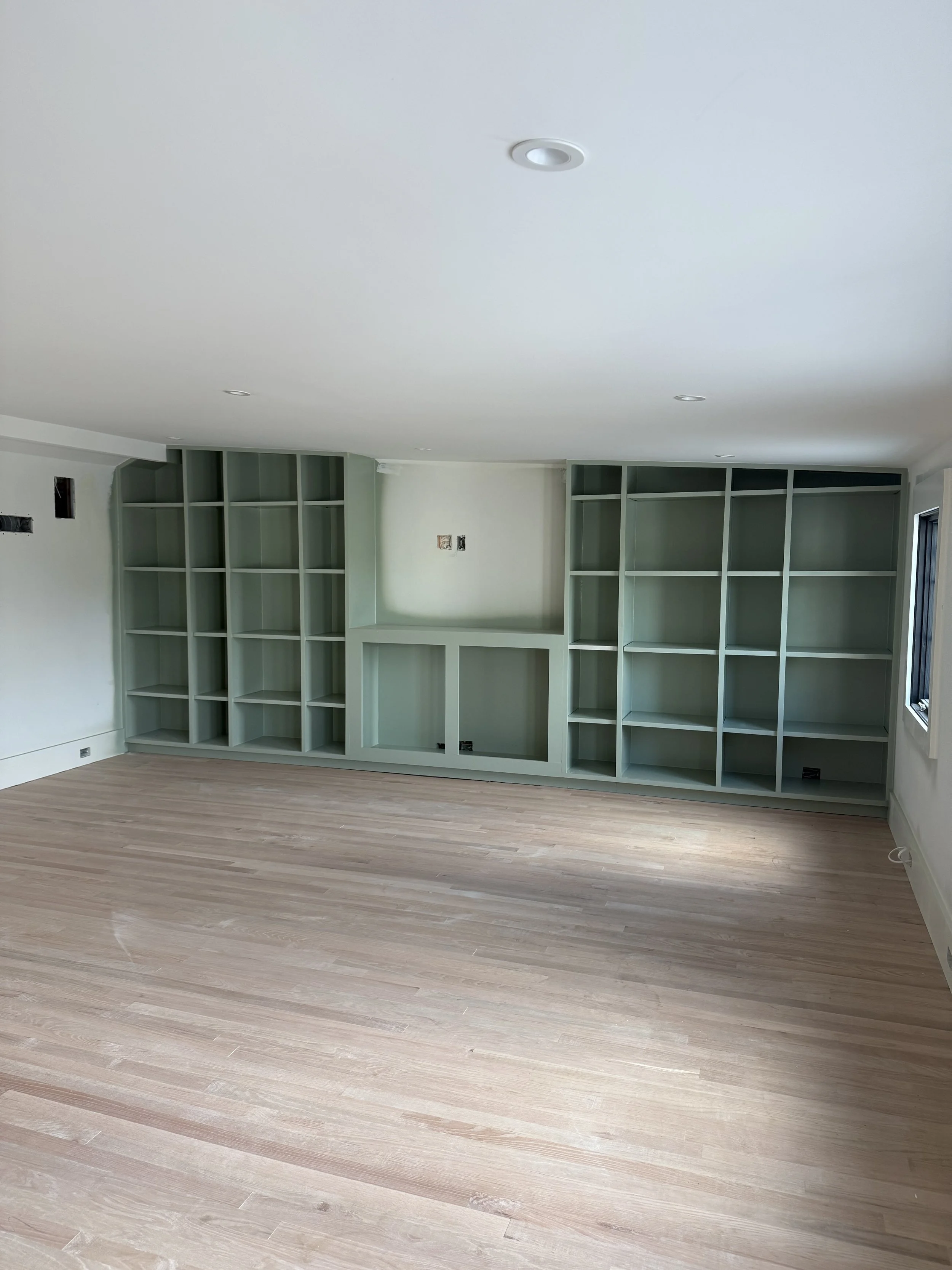 Empty living room with built-in green bookshelves on the wall, hardwood flooring, and a window on the right side.
