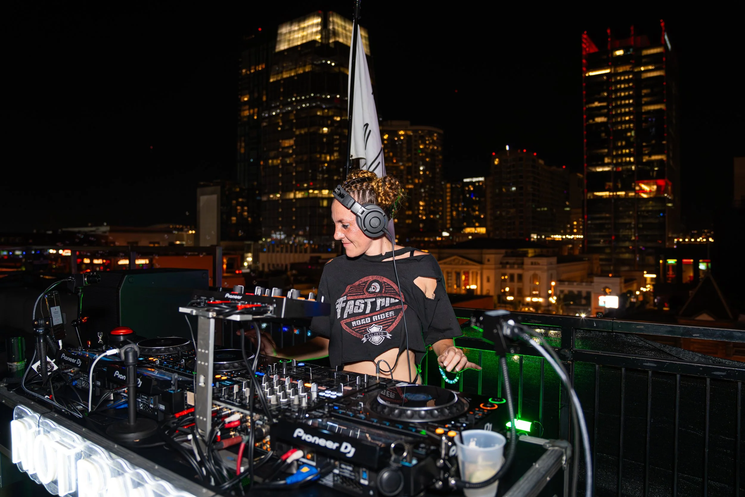 A woman DJ wearing headphones and peace sign earrings, working at a turntable and laptop, with sunlight filtering through a large window behind her.