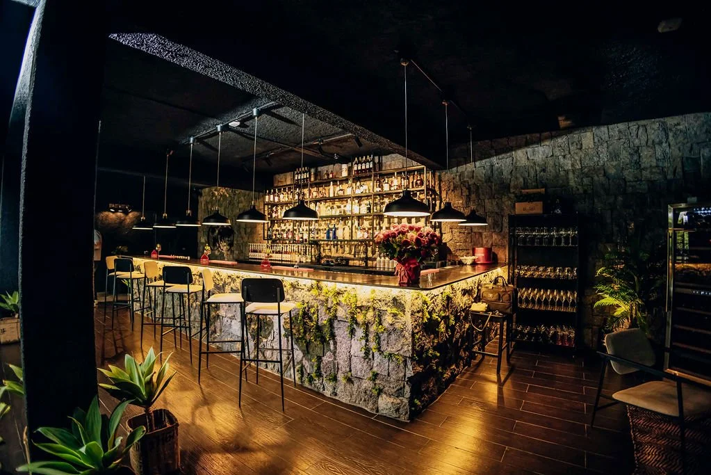 Dimly lit bar with a stone counter, hanging pendant lights, a flower bouquet on the counter, and shelves with alcohol bottles in the background. There are bar stools and potted plants around the space.