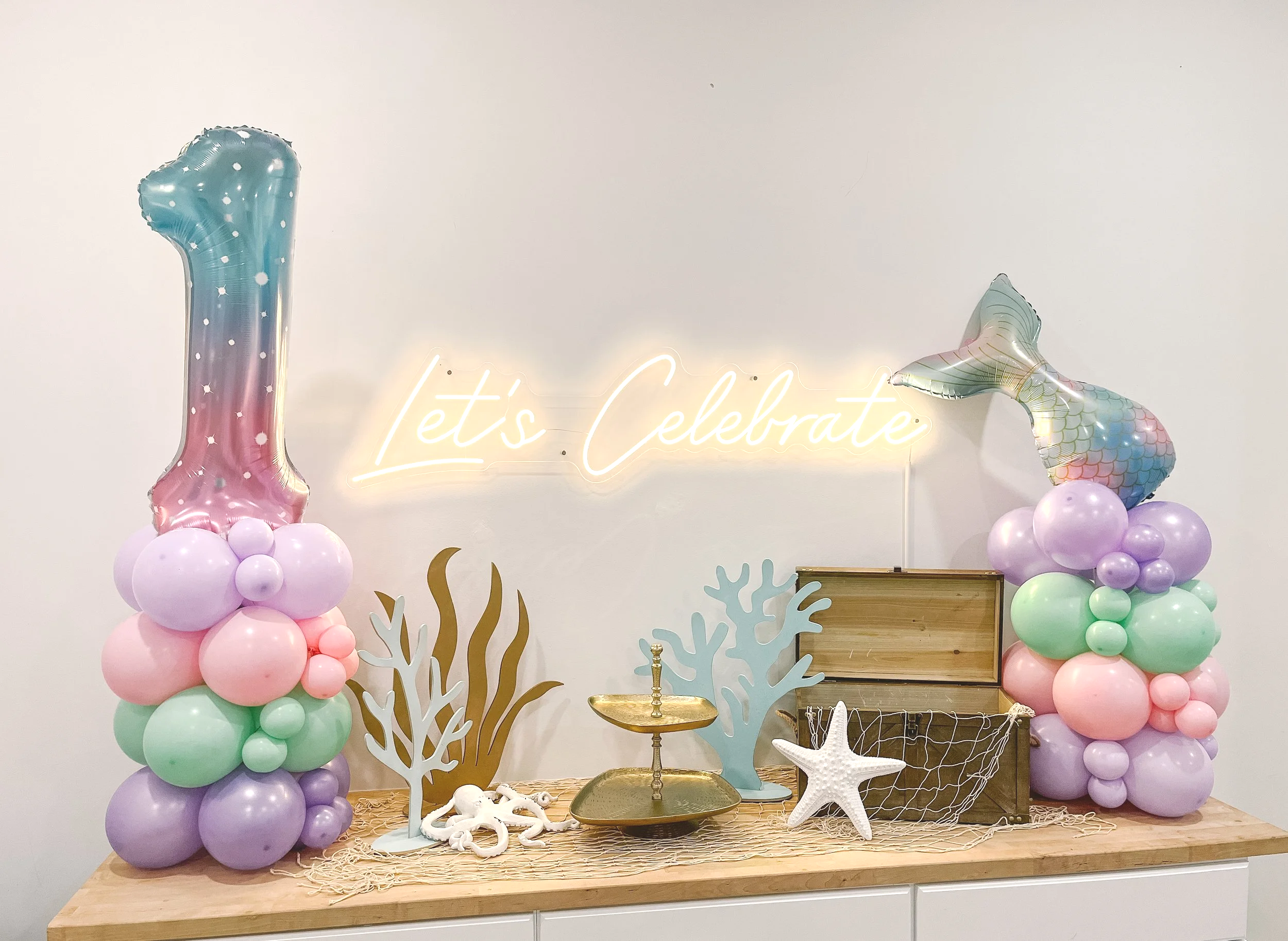 Decorative table with ocean-themed balloons, coral cutouts, starfish, seashells, and a neon sign that says 'Let's Celebrate' for a party or celebration.