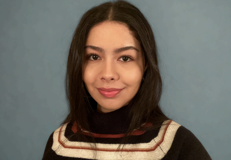 A young woman with shoulder-length dark hair and light brown skin, smiling slightly, wearing a black sweater with red, white, and beige stripes, indoors against a plain blue wall.
