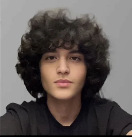 A person with curly, voluminous hair and a neutral expression, wearing a black shirt, against a plain gray background.