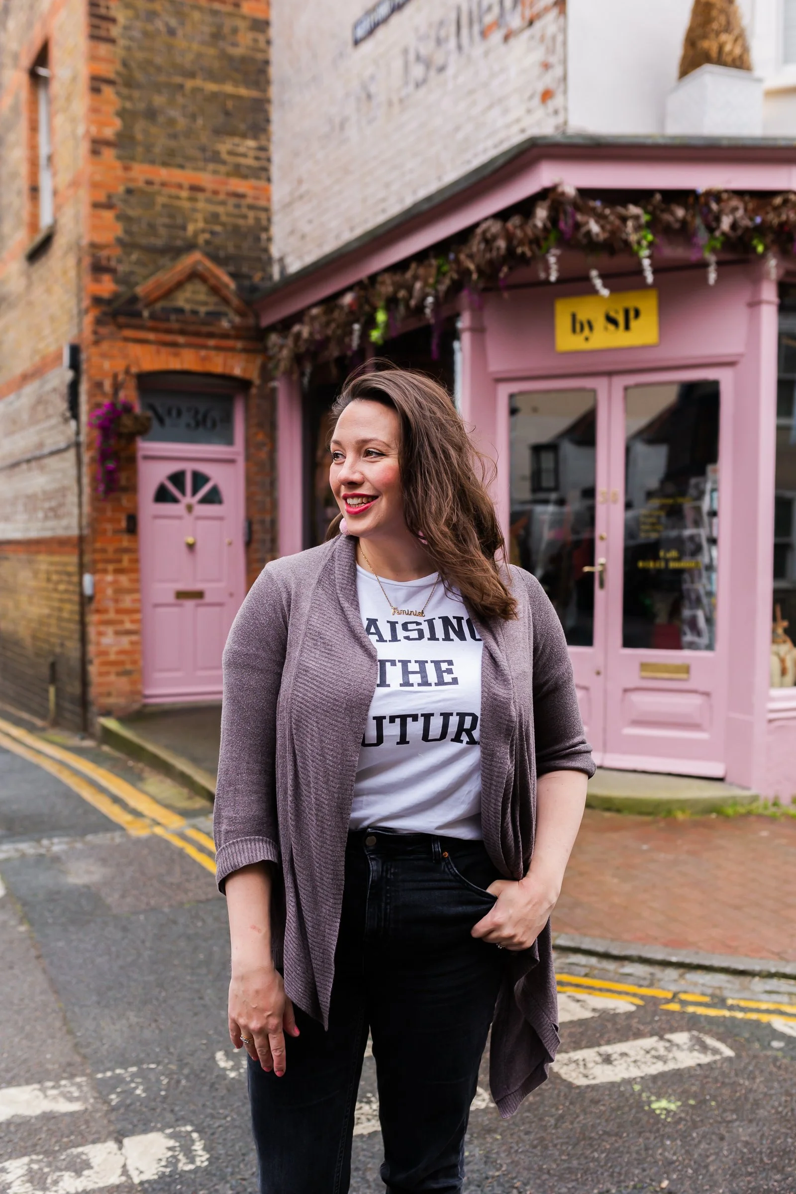 A woman with wavy brown hair, wearing a gray cardigan and white graphic T-shirt, smiling on a city street with a pink storefront and brick building behind her.